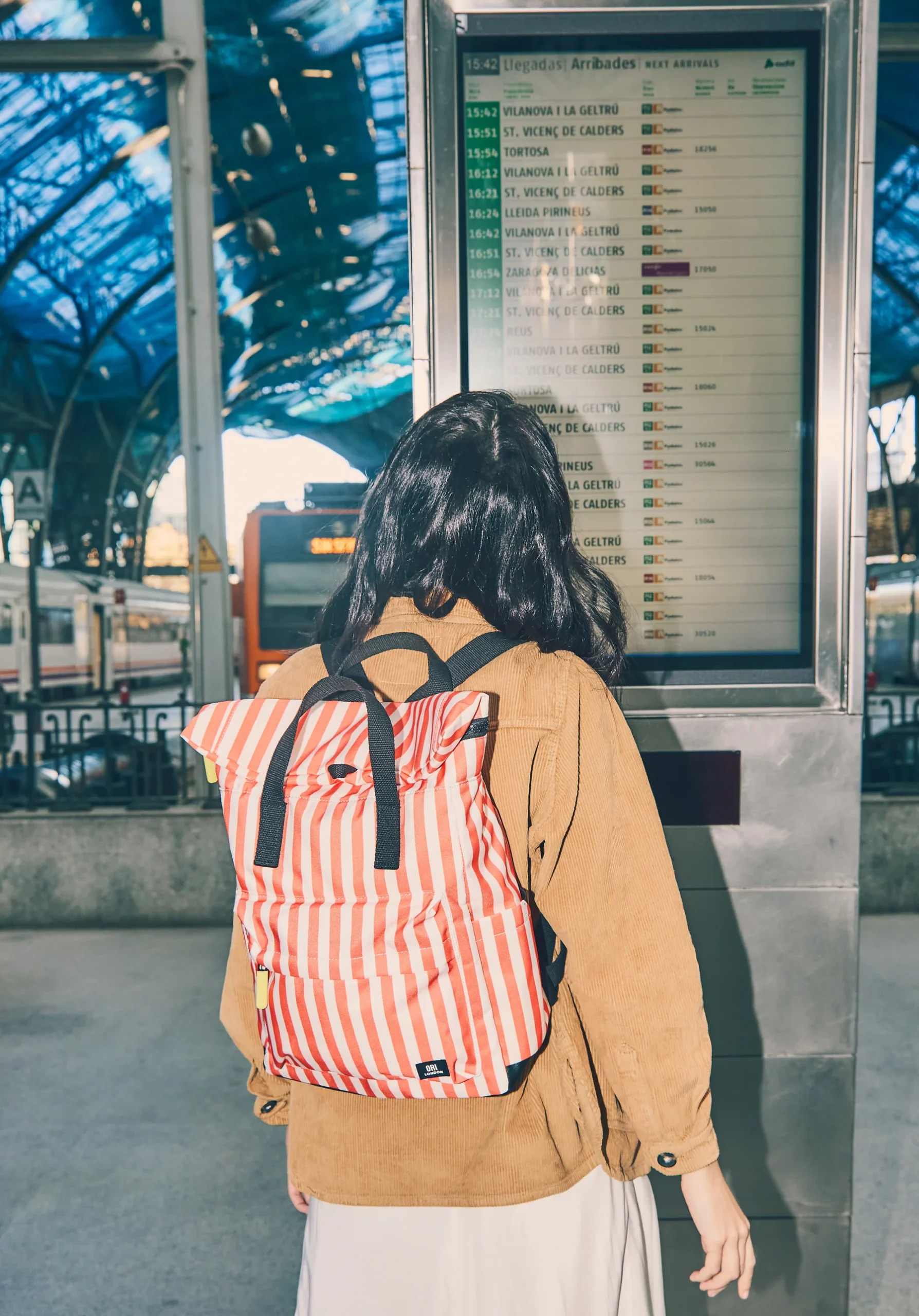 Person with long dark hair and a red striped backpack stands facing a digital arrivals board at a train station platform.