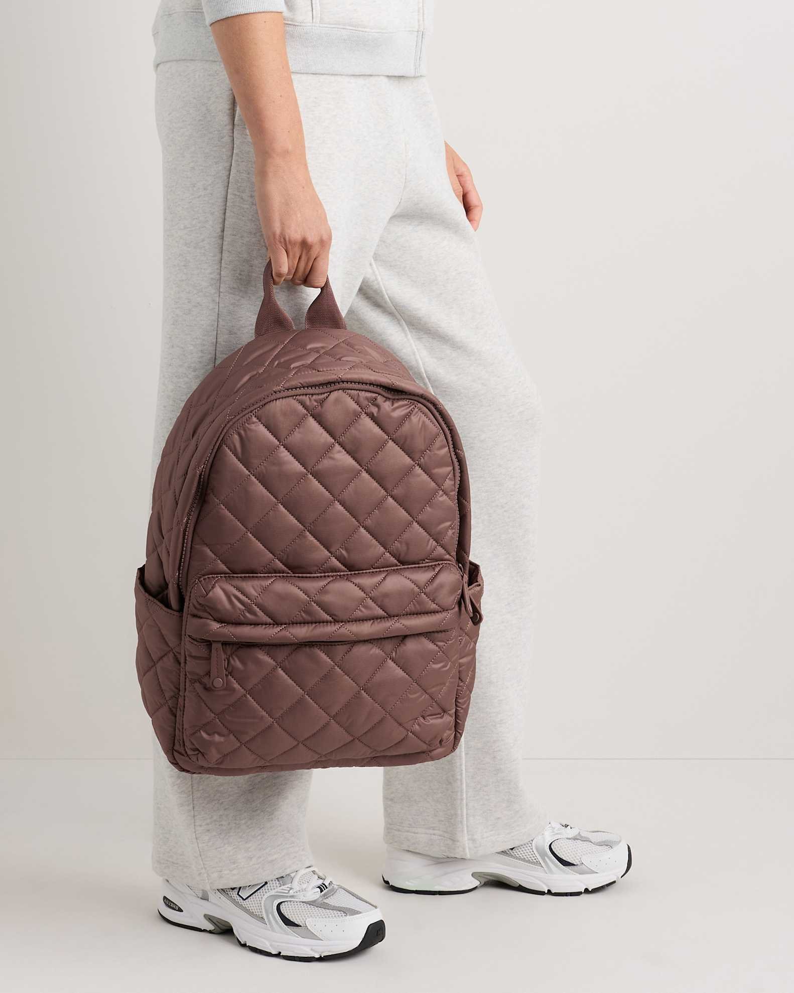 Person wearing light gray sweatpants and white sneakers holds a brown quilted backpack by the top handle, standing against a plain light background.