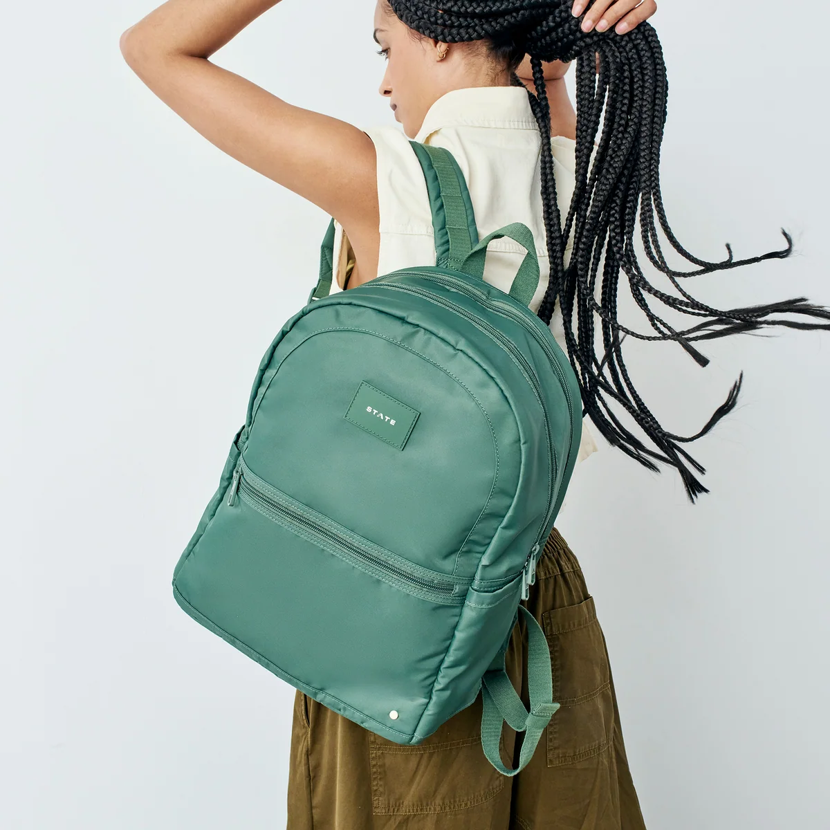 Person with long braids wearing a sleeveless shirt holds a large green backpack over one shoulder, facing away from the camera against a plain background.