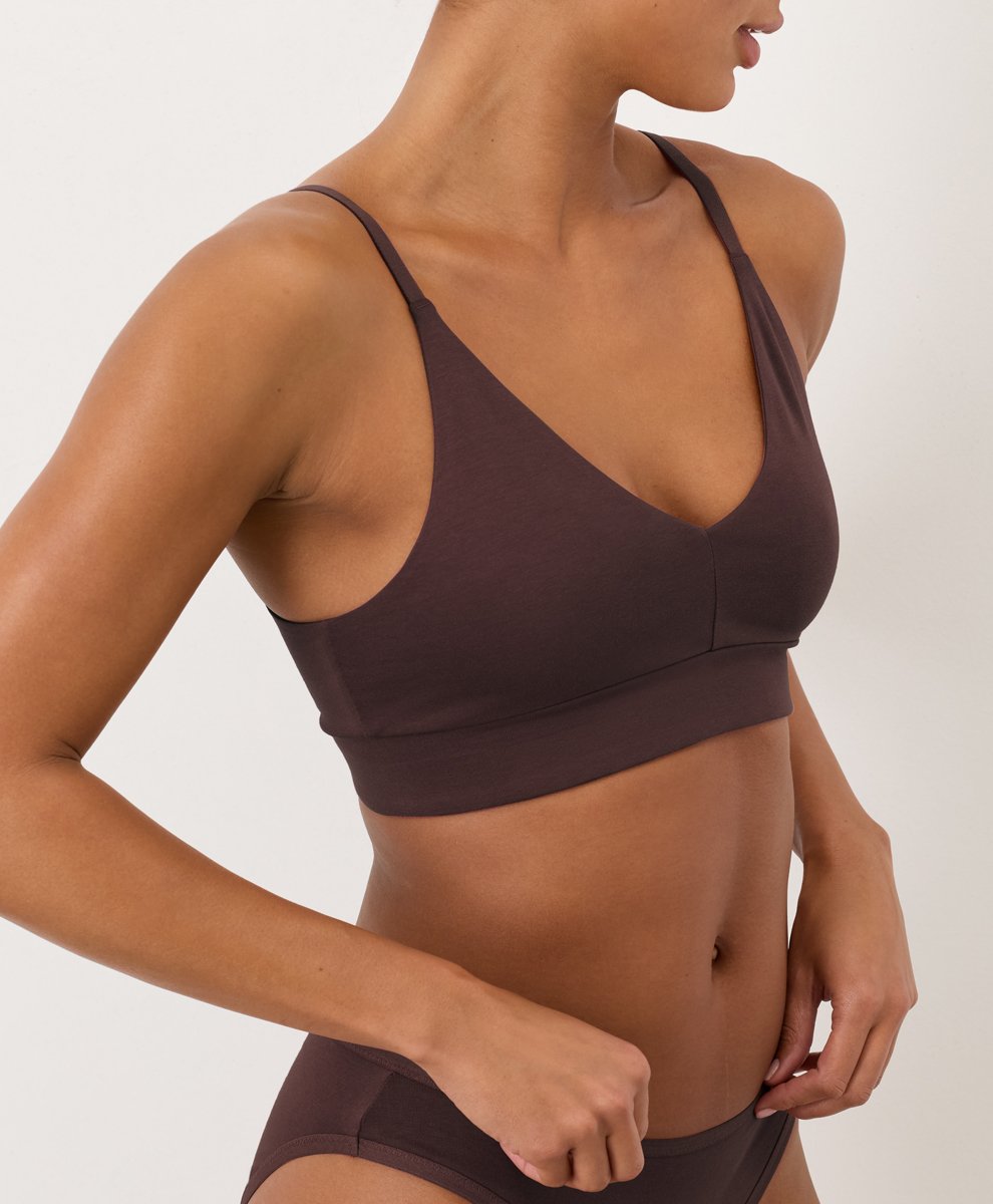 Person wearing a brown bralette and matching underwear, standing against a plain light background, with one arm bent and hand resting near the waistband.