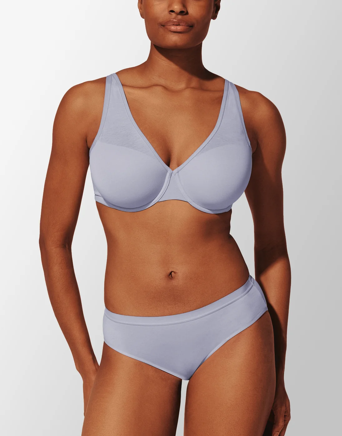 A woman stands facing forward wearing a matching light purple bra and underwear set against a plain, gradient background.