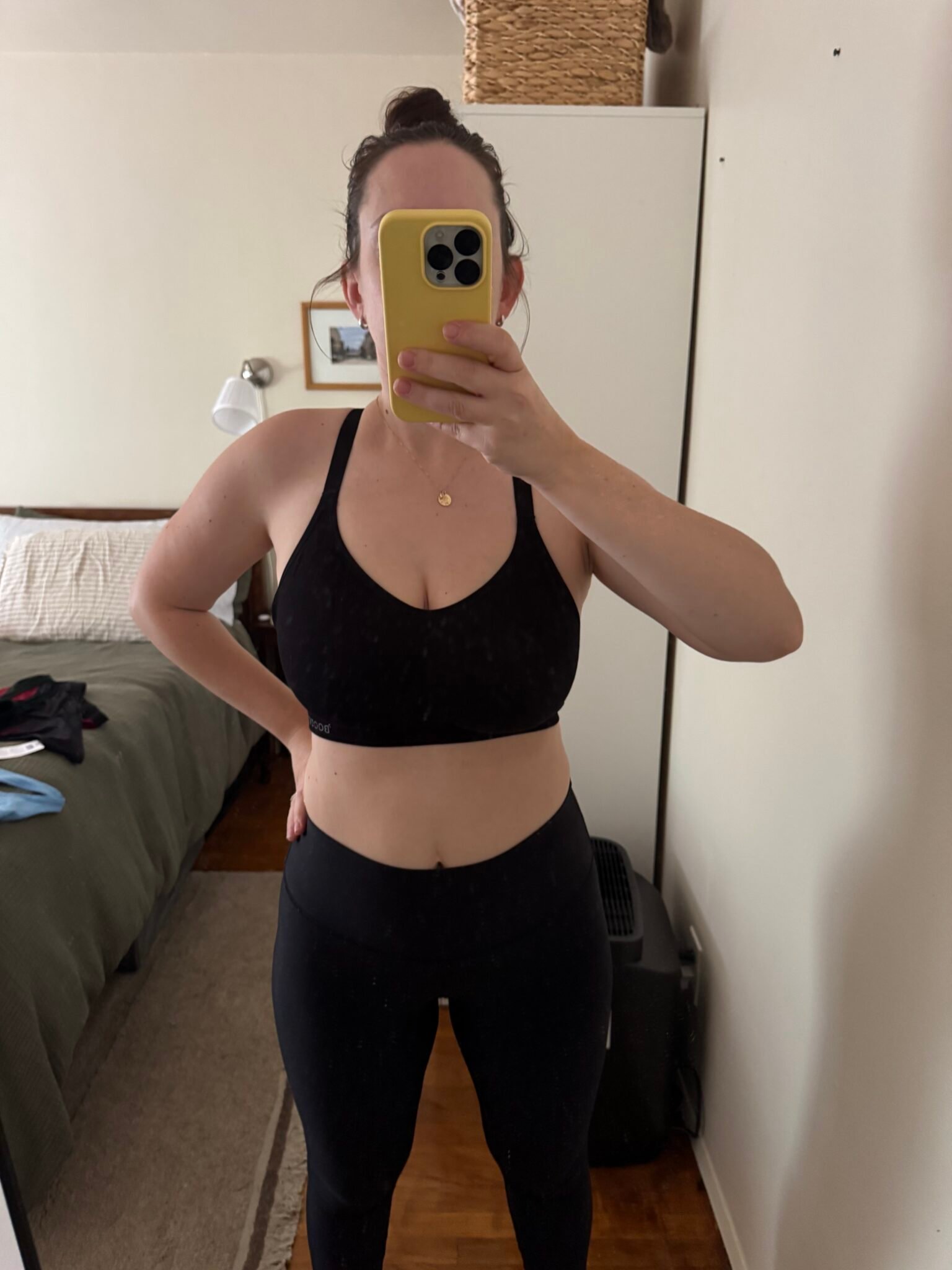 Person wearing a black sports bra and leggings takes a mirror selfie in a bedroom, partially obscuring their face with a yellow phone.