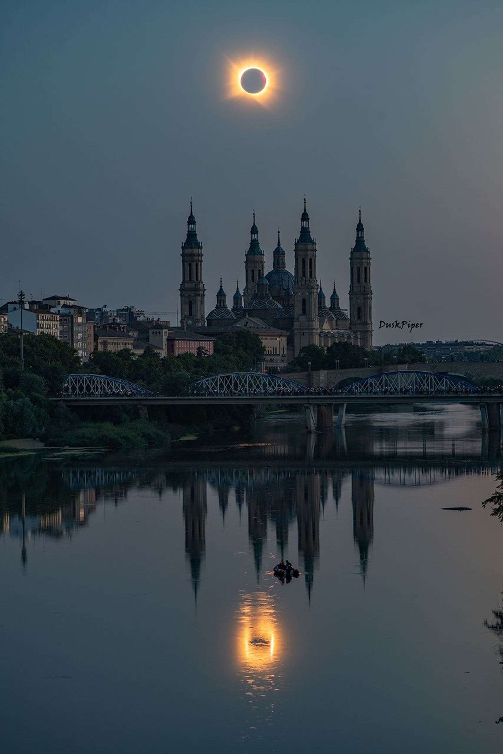 The Moon covers the Sun in the middle of the sky above an ornate church. A ring of bright whisps from the Sun's corona surrounds the Moon. In front of the church is a river that shows ripples across the eclipse's reflection. Some people are boating in the river.