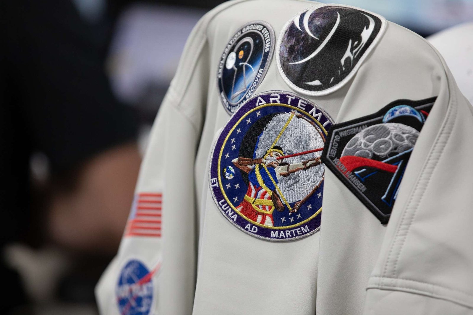 A gray colored jacket with five Artemis mission patches and a NASA insignia hangs on a chair.