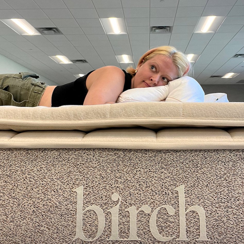 A person relaxes on a mattress with their head on a pillow. The word "birch" graces the side of this nontoxic, organic mattress. The room features a ceiling adorned with lights and vents, setting the perfect scene for a glowing mattress review.