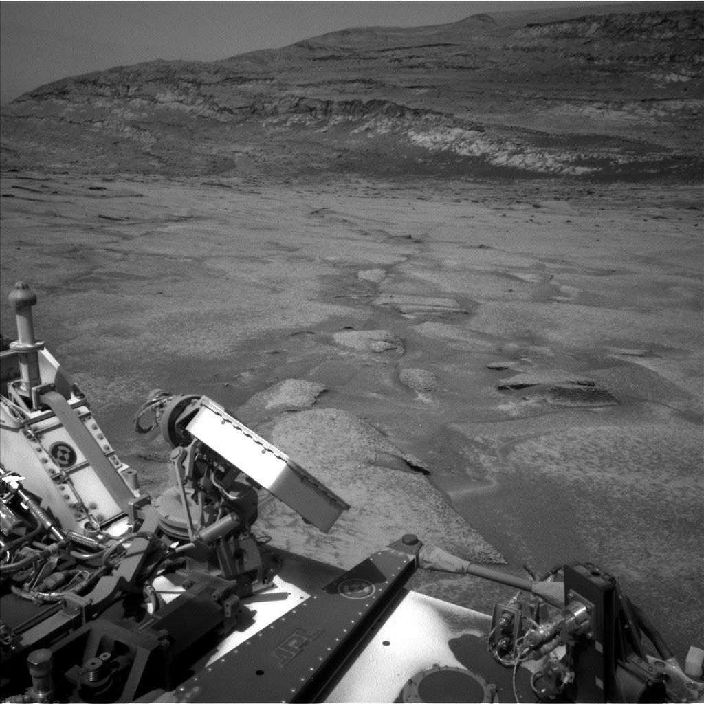 A black and white image taken from the deck of the Curiosity rover on Mars, looking out over a desolate, rocky landscape. In the foreground, parts of the rover's hardware, including mechanical joints, structural panels, and a dark strut bearing the 'JPL' logo, are visible. The middle ground features relatively flat terrain covered in dusty soil, dotted with light-colored, exposed rock plates and shallow depressions. In the background, rolling hills or mountains rise, displaying distinct, horizontal sedimentary layers against a clear, featureless sky.
