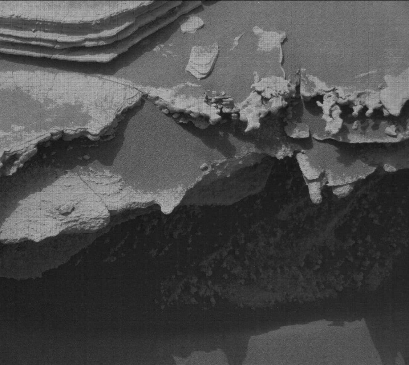A black-and-white close-up from the Curiosity rover showing a jagged, deeply eroded rock ledge casting a dark shadow, with thinly layered, flat sedimentary rocks visible in the upper left corner.