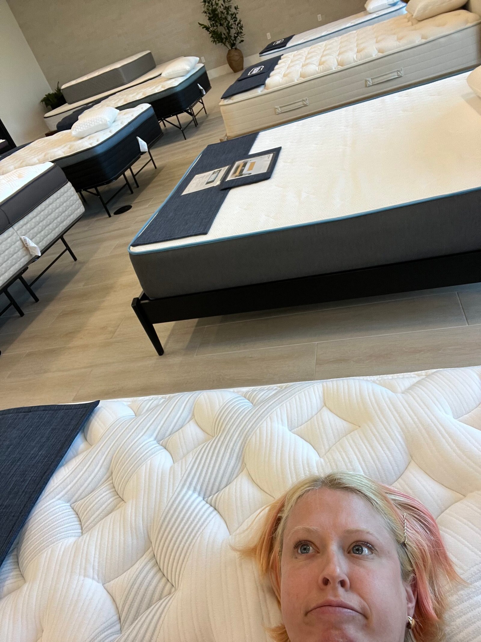 Person lying on a mattress in a showroom filled with neatly arranged mattresses on display.