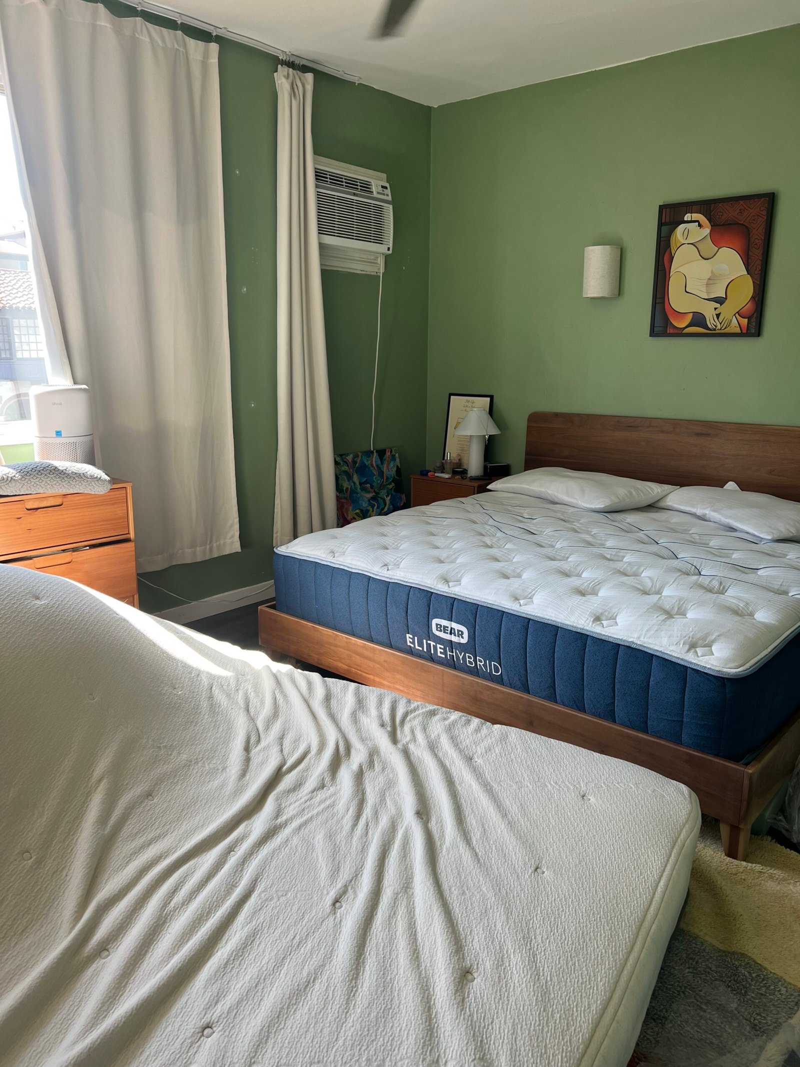 Bedroom with green walls featuring two beds, one with a blue mattress labeled "Elite Hybrid" and the other bare. The room has a wall-mounted air conditioner, white curtains, and a framed painting.