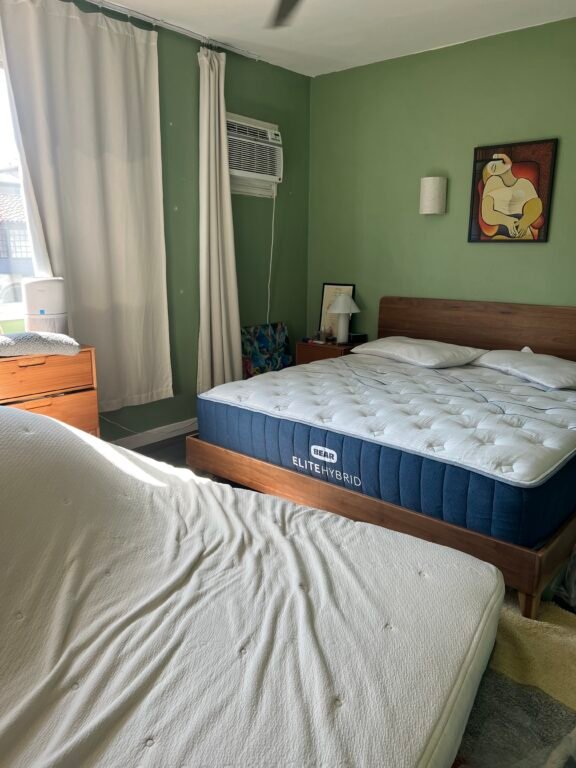 A bedroom with two beds, one upright and one at an angle, green walls, a window with a curtain, an air conditioner, a nightstand, and a colorful painting on the wall.