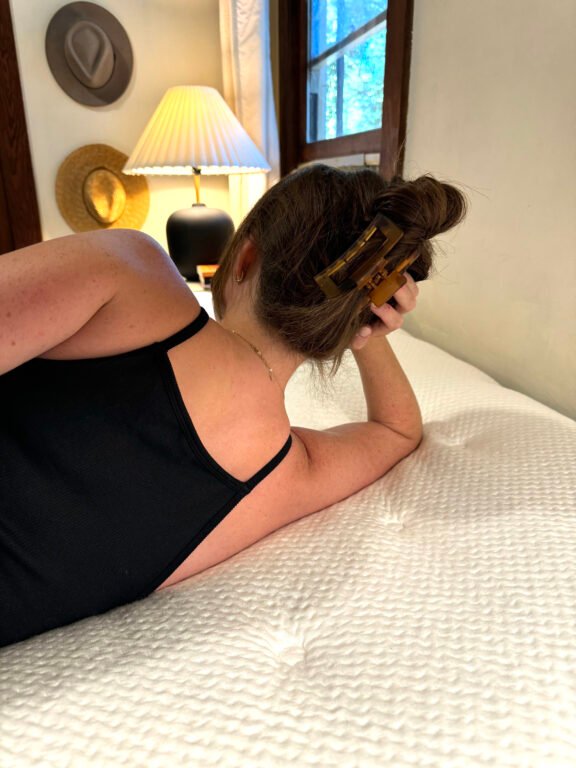 A person with a hair clip lies on their side on a textured white mattress in a room with hats on the wall and a lamp on a bedside table.
