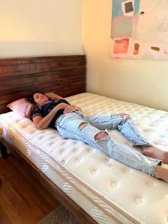 A woman in a black t-shirt and ripped jeans lies on her back on a bare mattress in a bedroom with wooden floors and abstract art on the wall.