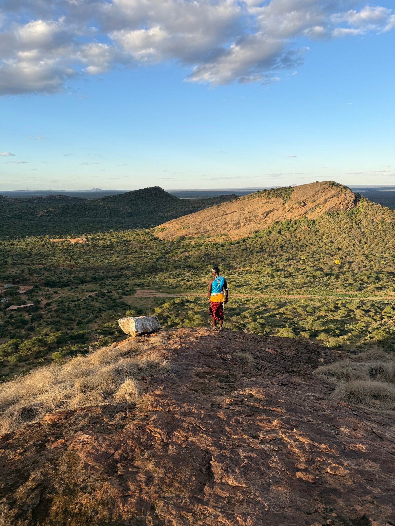 A person in colorful clothing stands on a rocky hilltop overlooking a vast, green landscape with distant hills under a partly cloudy sky.