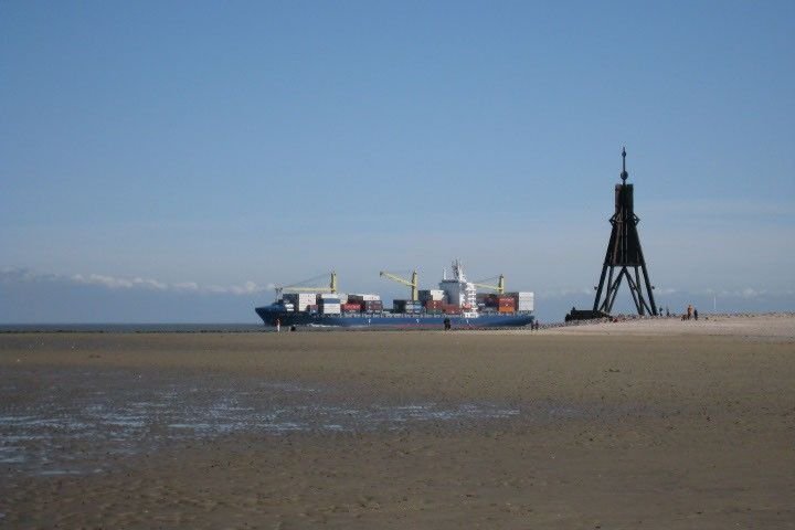 A broad tidal mudflat fills the foreground of this photo. A sliver of deeper water is visible on the horizon, where a cargo ship loaded with shipping containers and cranes appears to be traveling to the left.