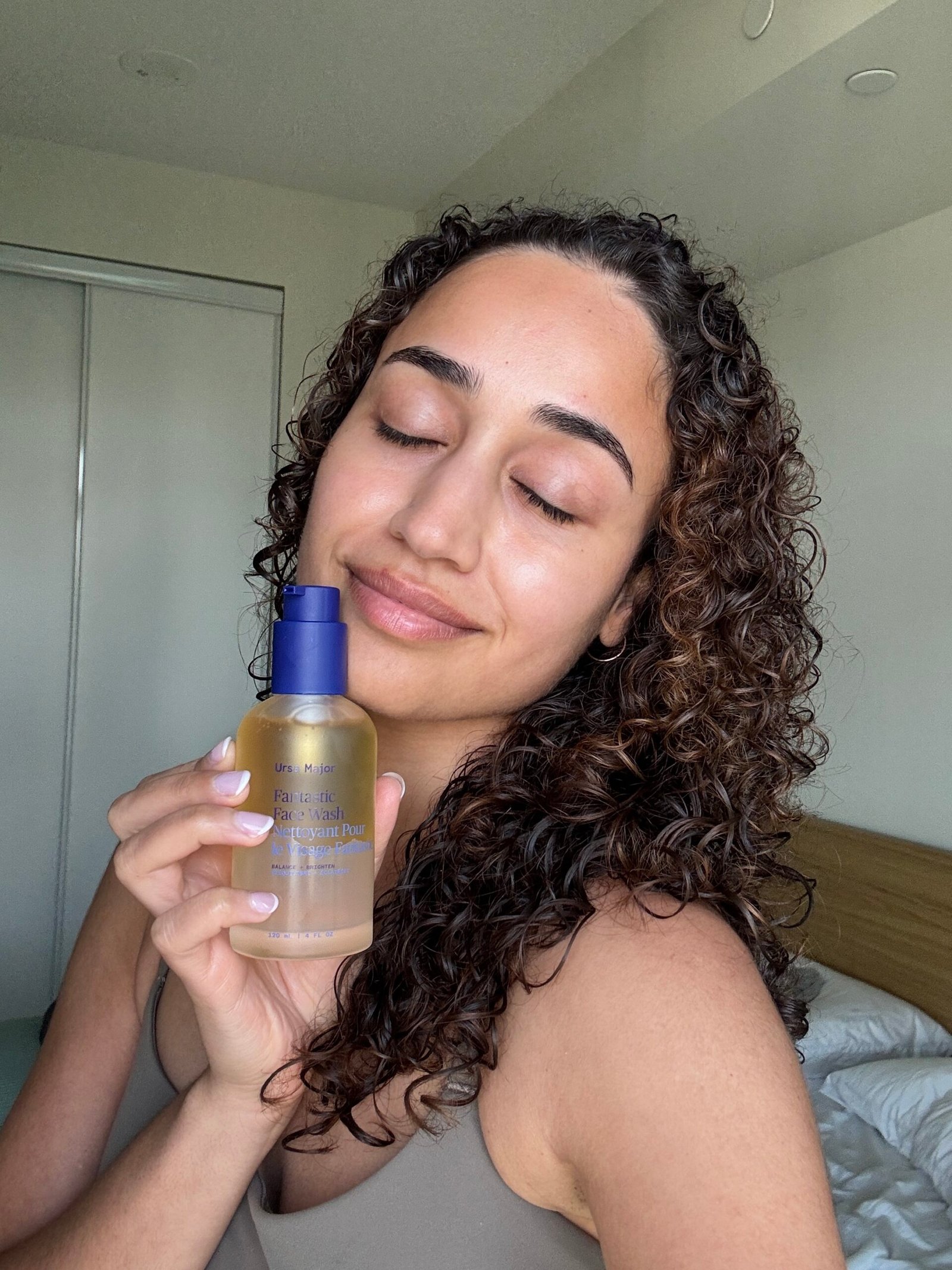 Person with curly hair holds a translucent bottle of skincare product, eyes closed, smiling slightly, standing indoors near a bed and closet.