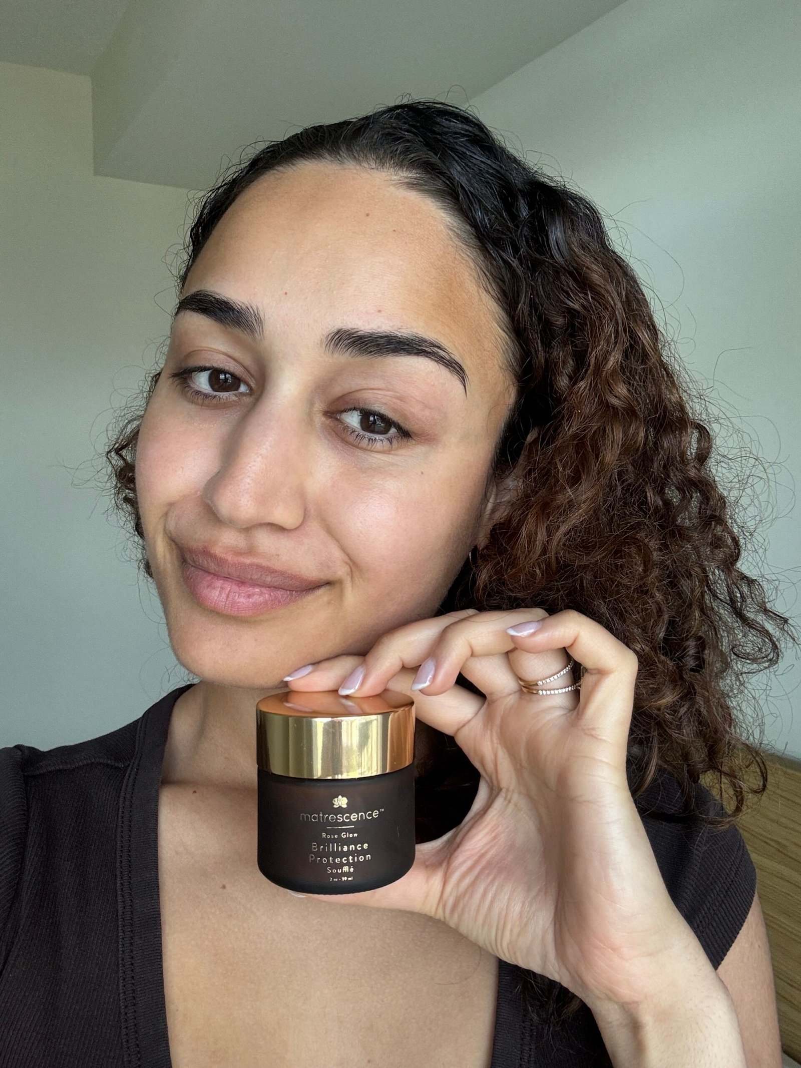 A person with curly hair holds a jar of Matrescence skincare product close to their face, smiling softly at the camera.