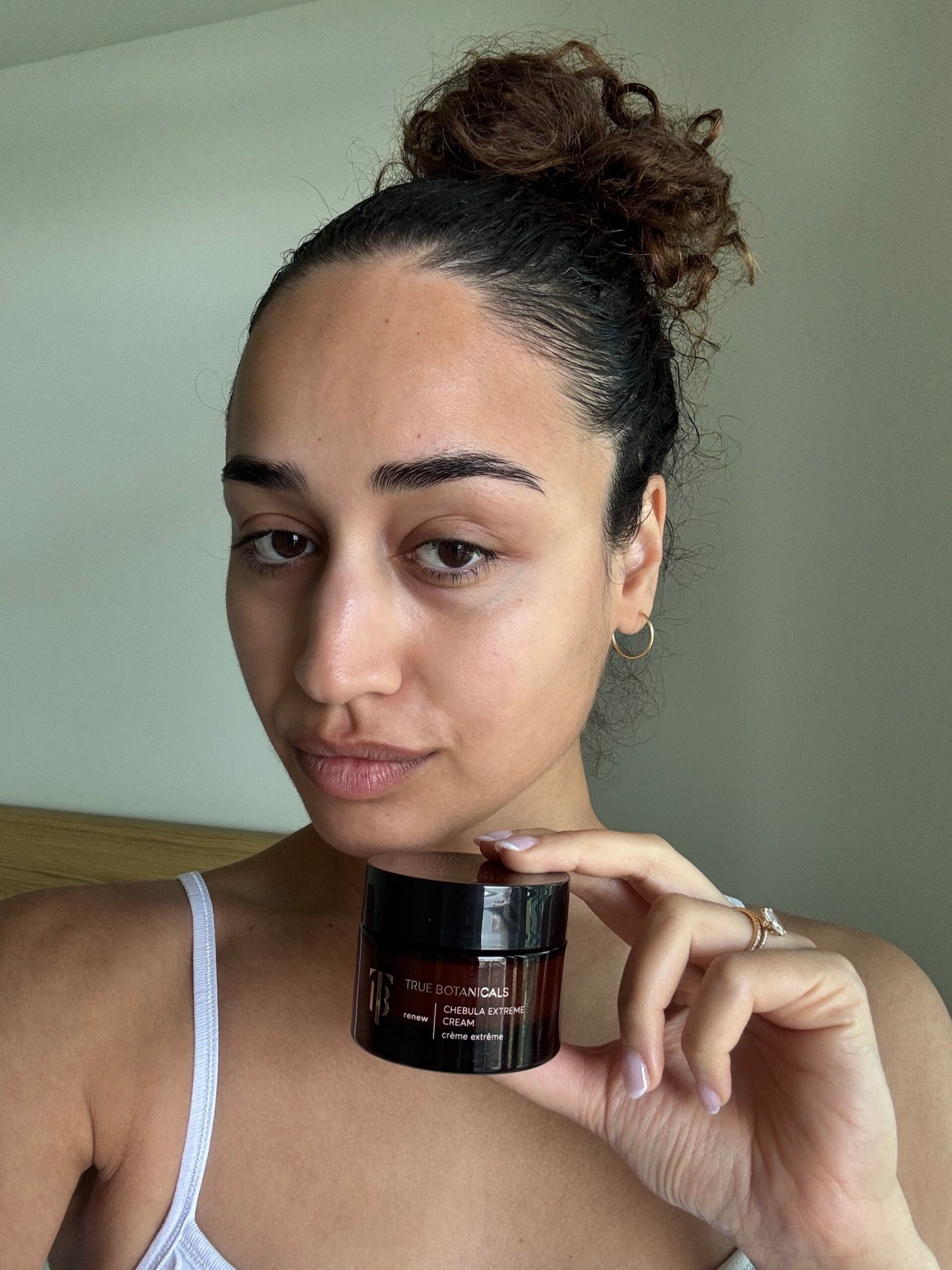 Woman with pulled-back hair and minimal makeup holds a jar of True Botanicals Chebula Extreme Cream, standing indoors with a neutral background.