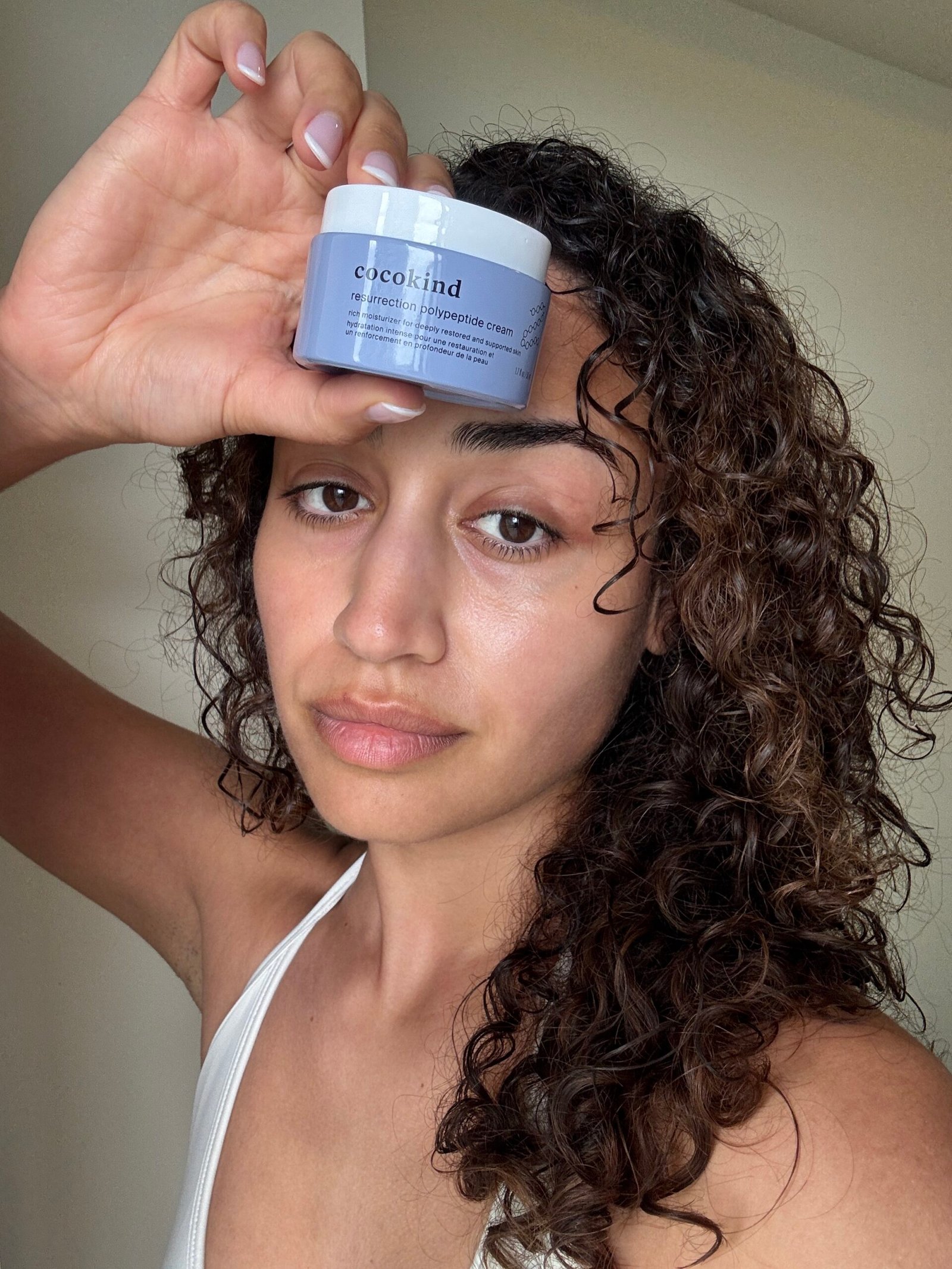 Person with curly hair holds a jar of Cocokind Resurrection Polypeptide Cream next to their face, wearing a white top, in a well-lit indoor setting.