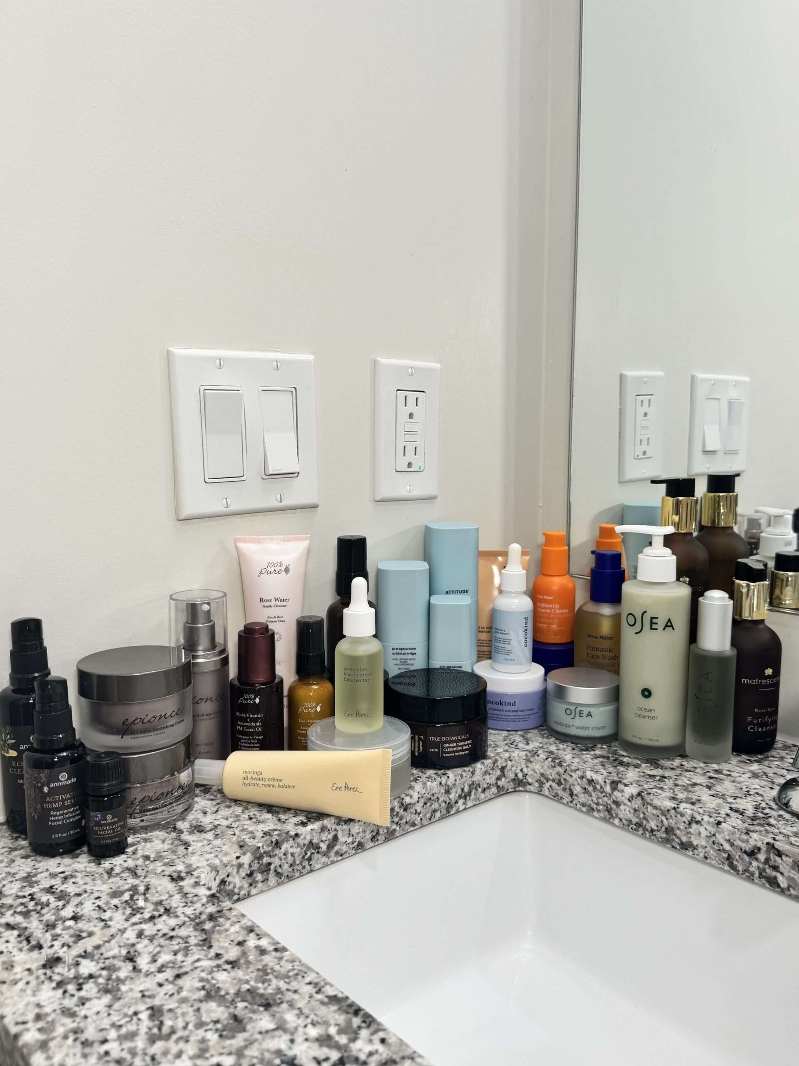 A variety of skincare and beauty products are arranged on a granite bathroom countertop next to a sink and mirror.