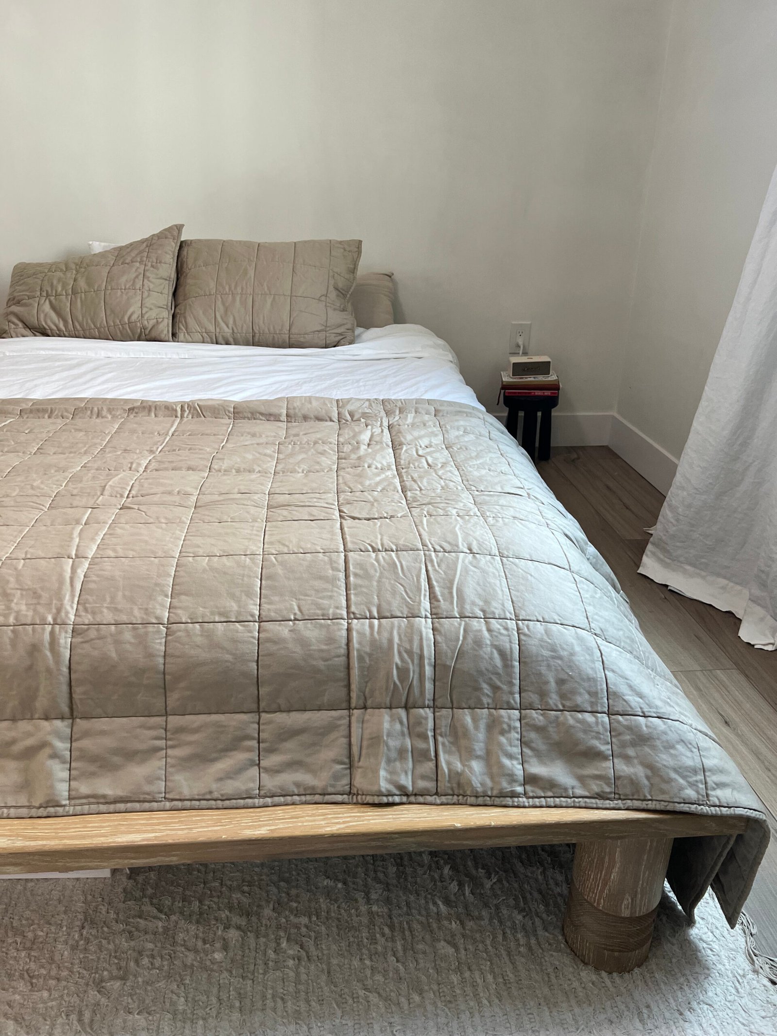 A neatly made bed with a beige quilt, matching pillows, and wooden bed frame. A small nightstand with a book and lamp is visible next to the bed. A white curtain hangs on the right.