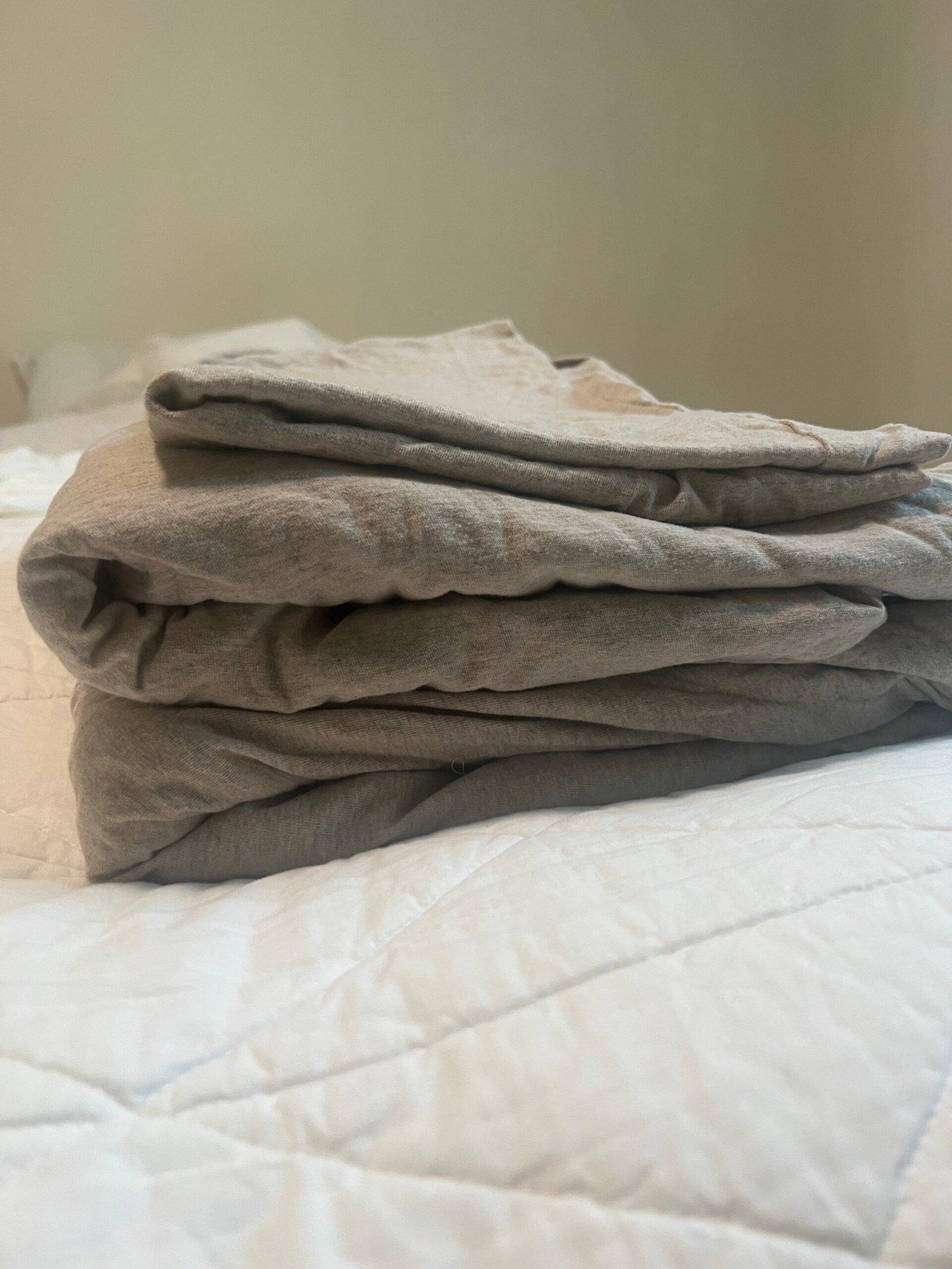 A neatly folded beige blanket sits on a white quilted bedspread in a softly lit room.