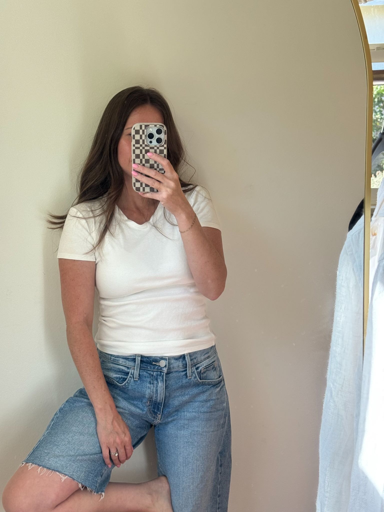 Woman in a white t-shirt and denim shorts takes a mirror selfie against a plain wall, holding a phone with a checkered case.