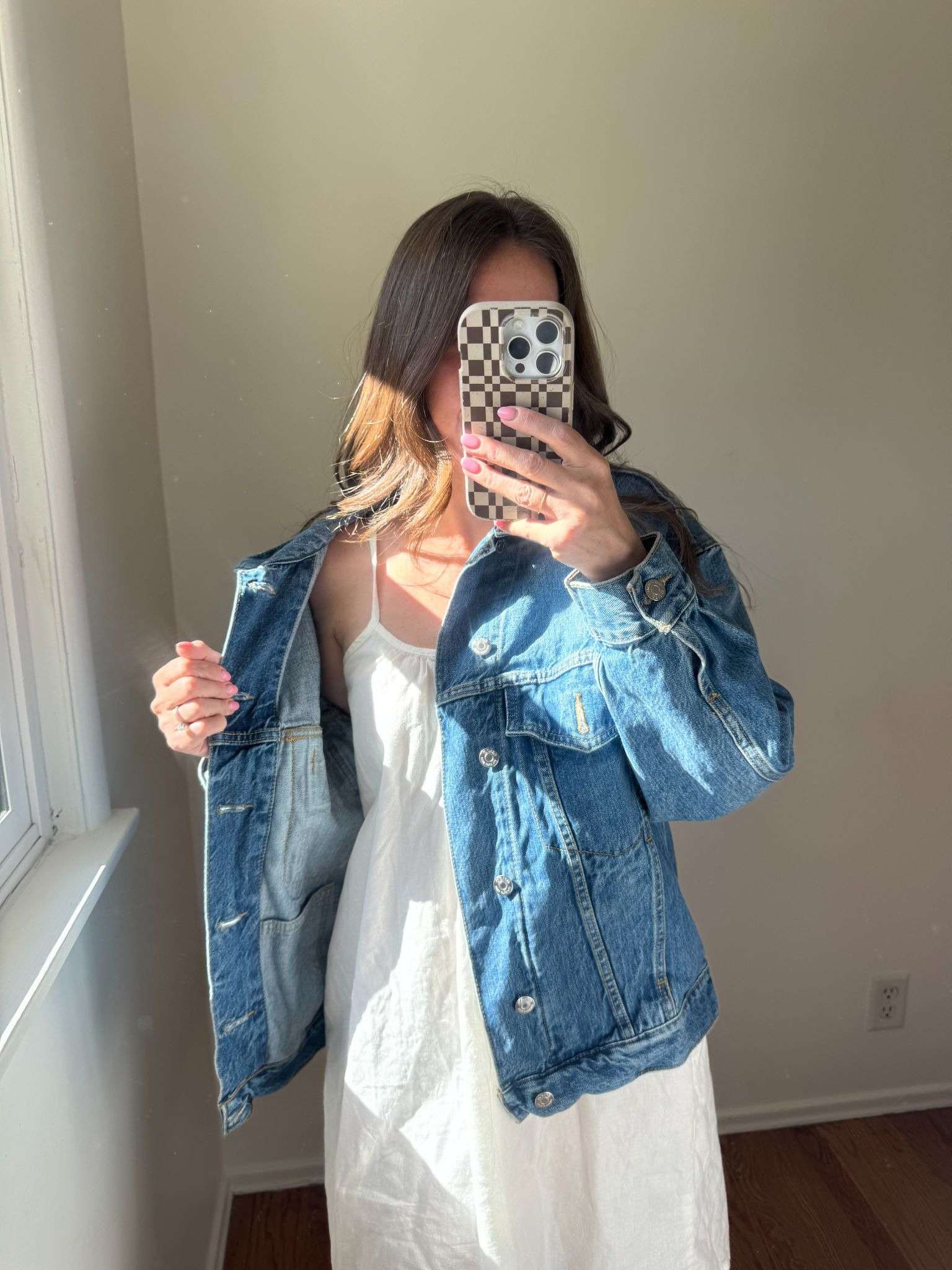 Person wearing a white dress and blue denim jacket stands indoors, taking a mirror selfie with a checkered phone case. Sunlight enters through a nearby window.