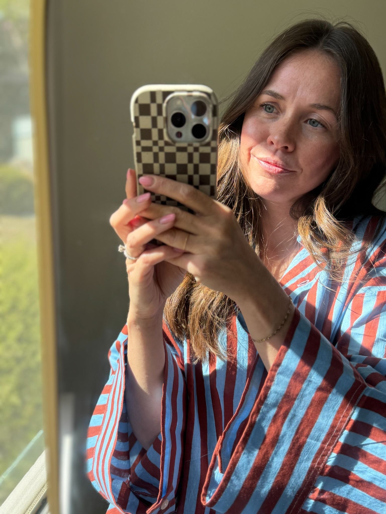 A woman in a blue and brown striped robe takes a selfie with a checker-patterned phone by a sunlit window.