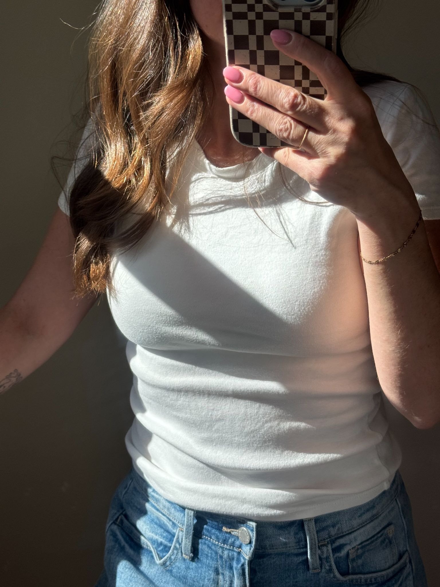 A person in a white t-shirt and blue jeans takes a mirror selfie using a phone with a checkered case. Sunlight highlights part of their torso and hair.