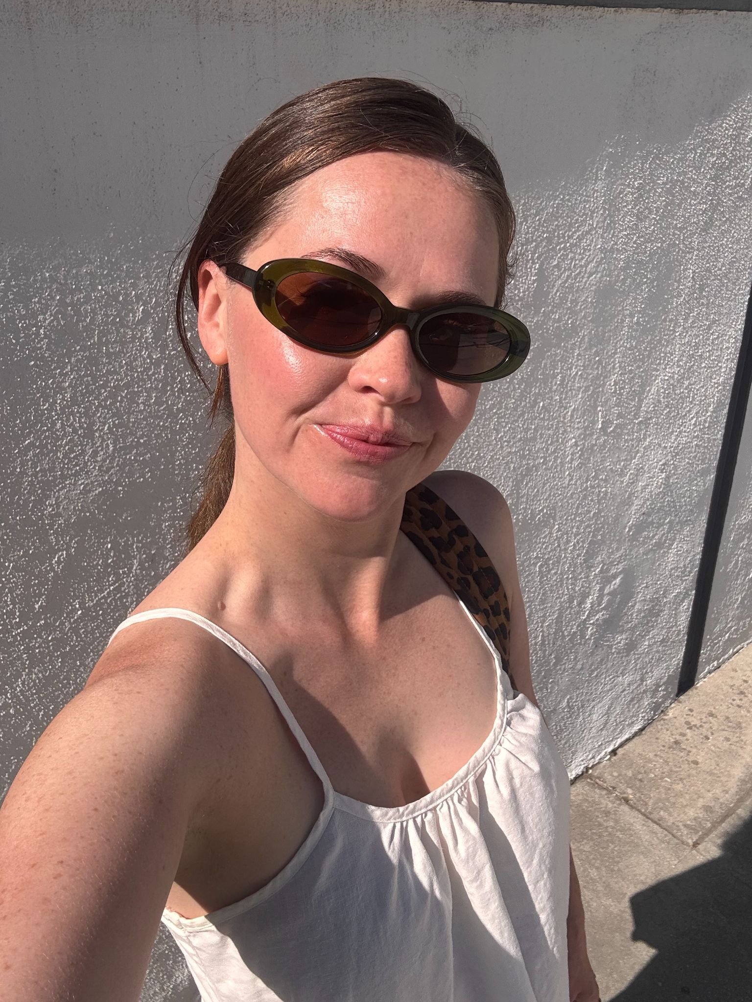 Woman wearing sunglasses and a white tank top stands outside in bright sunlight, taking a selfie against a white textured wall.
