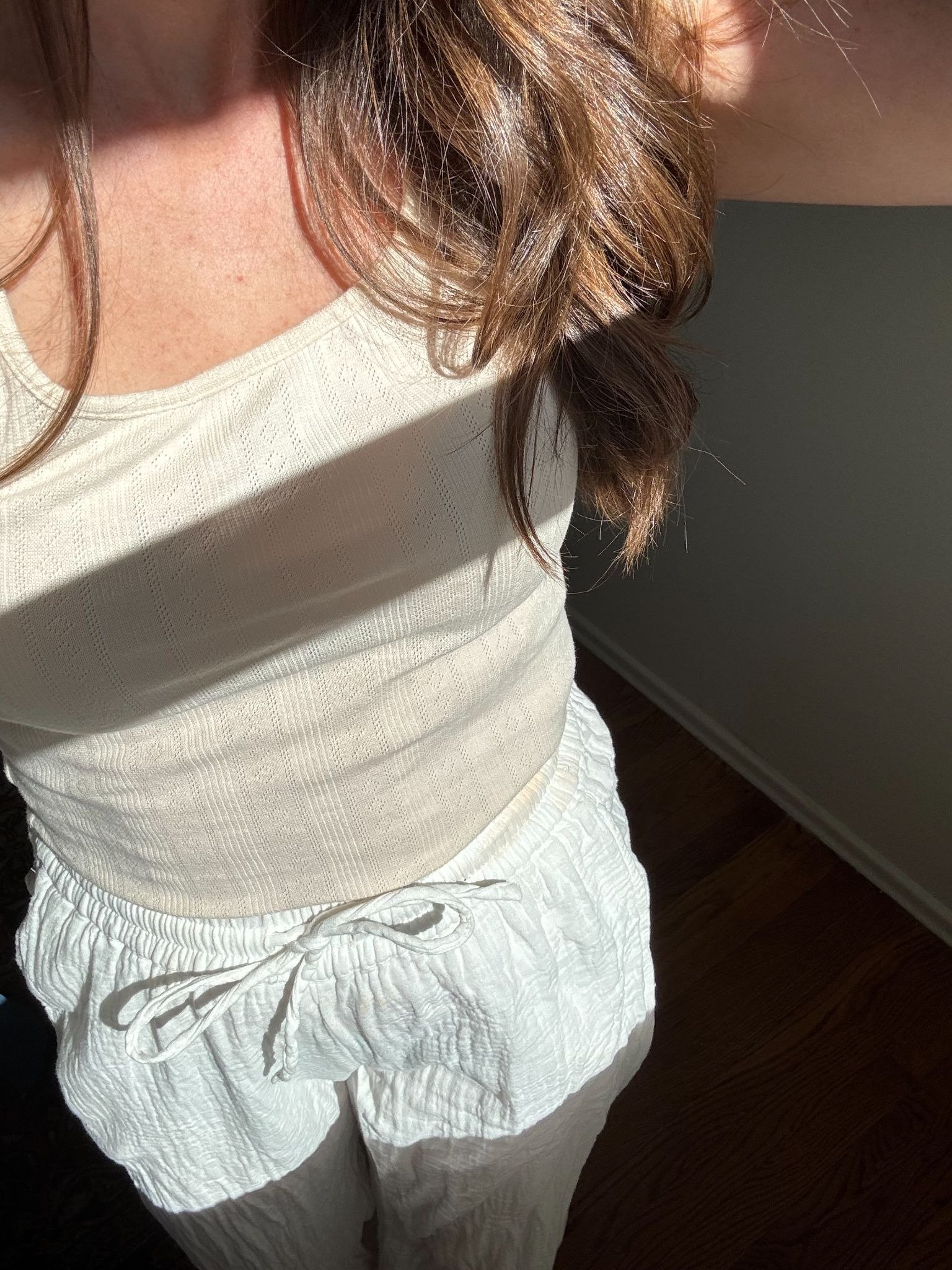 Woman wearing a cream-colored tank top and drawstring pants stands in sunlight, with her face out of frame and brown hair cascading over her shoulder.