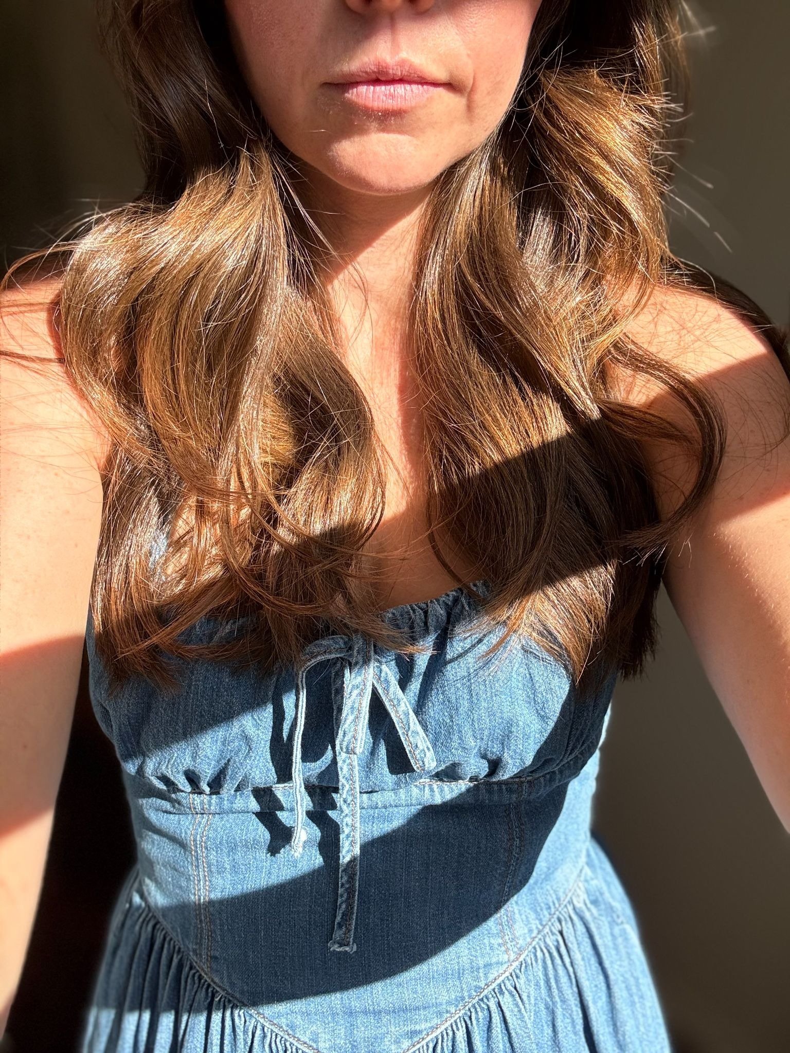 A woman wearing a blue denim dress is standing in sunlight, with her face partially visible and long wavy brown hair over her shoulders.