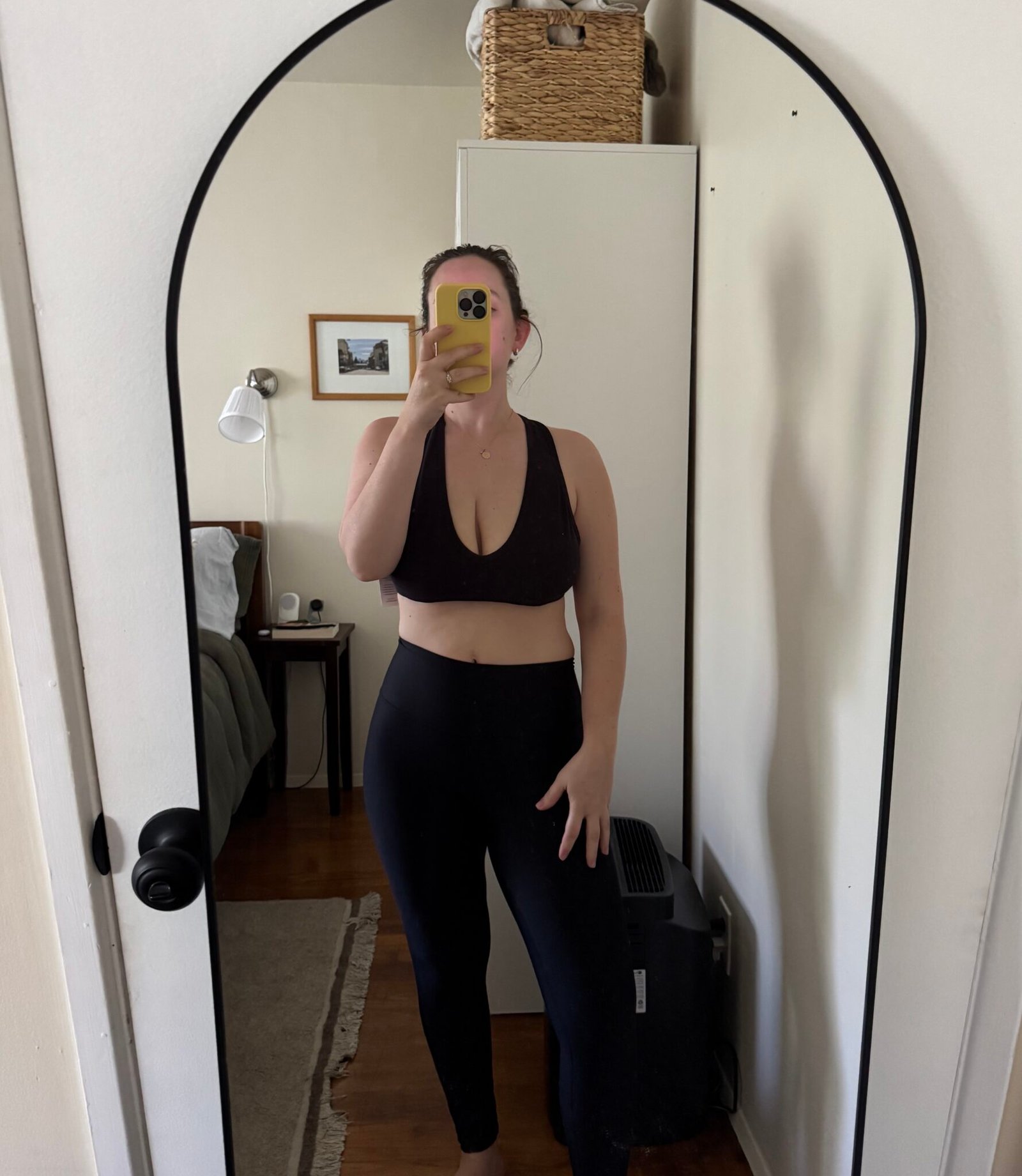 A person wearing a black sports bra and black leggings takes a mirror selfie in a bedroom with a bed, nightstand, and wall art visible in the background.