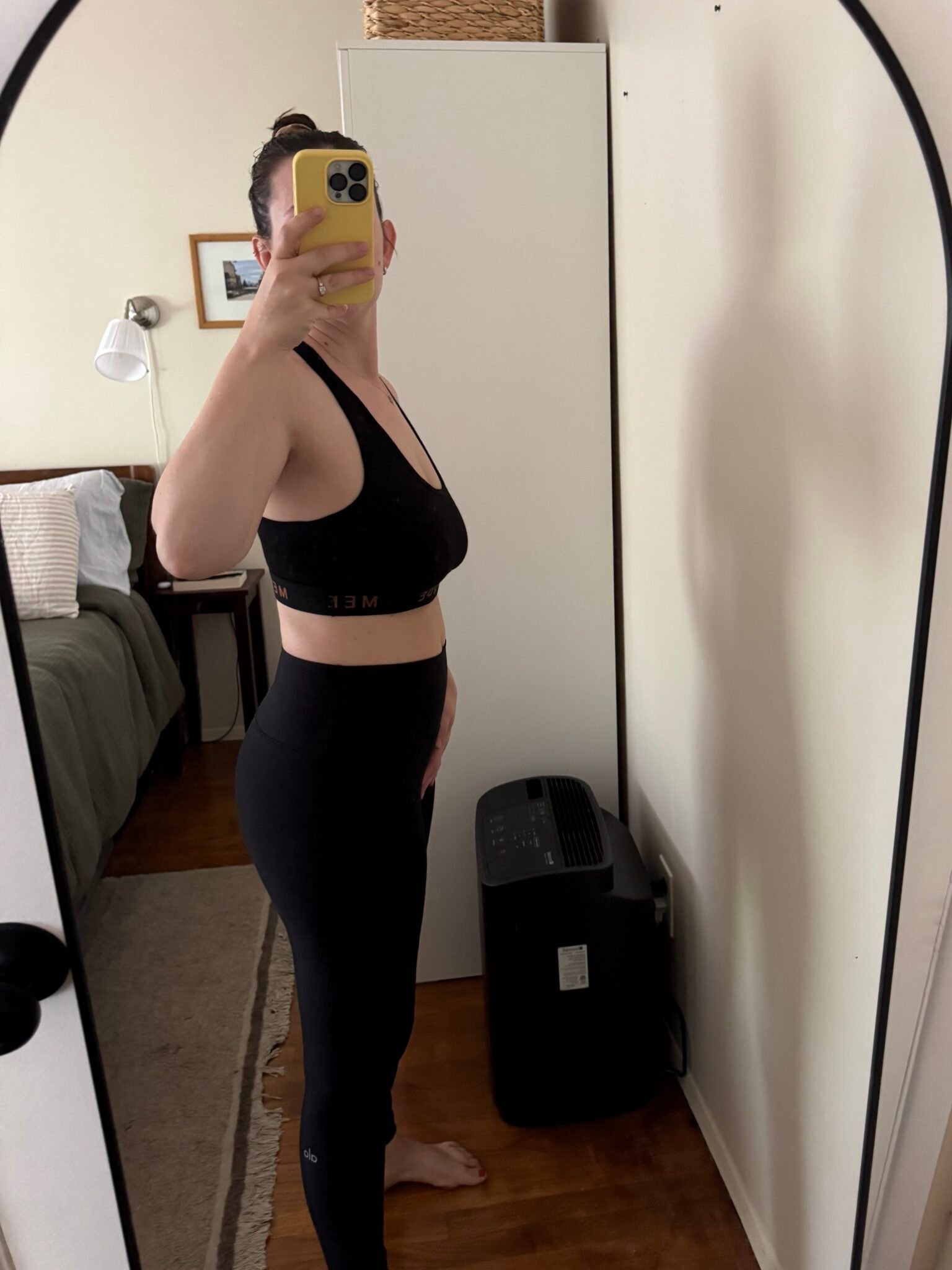 Person wearing black athletic wear stands sideways in front of a mirror, taking a photo with a yellow phone in a bedroom with a white cabinet and a bed visible.