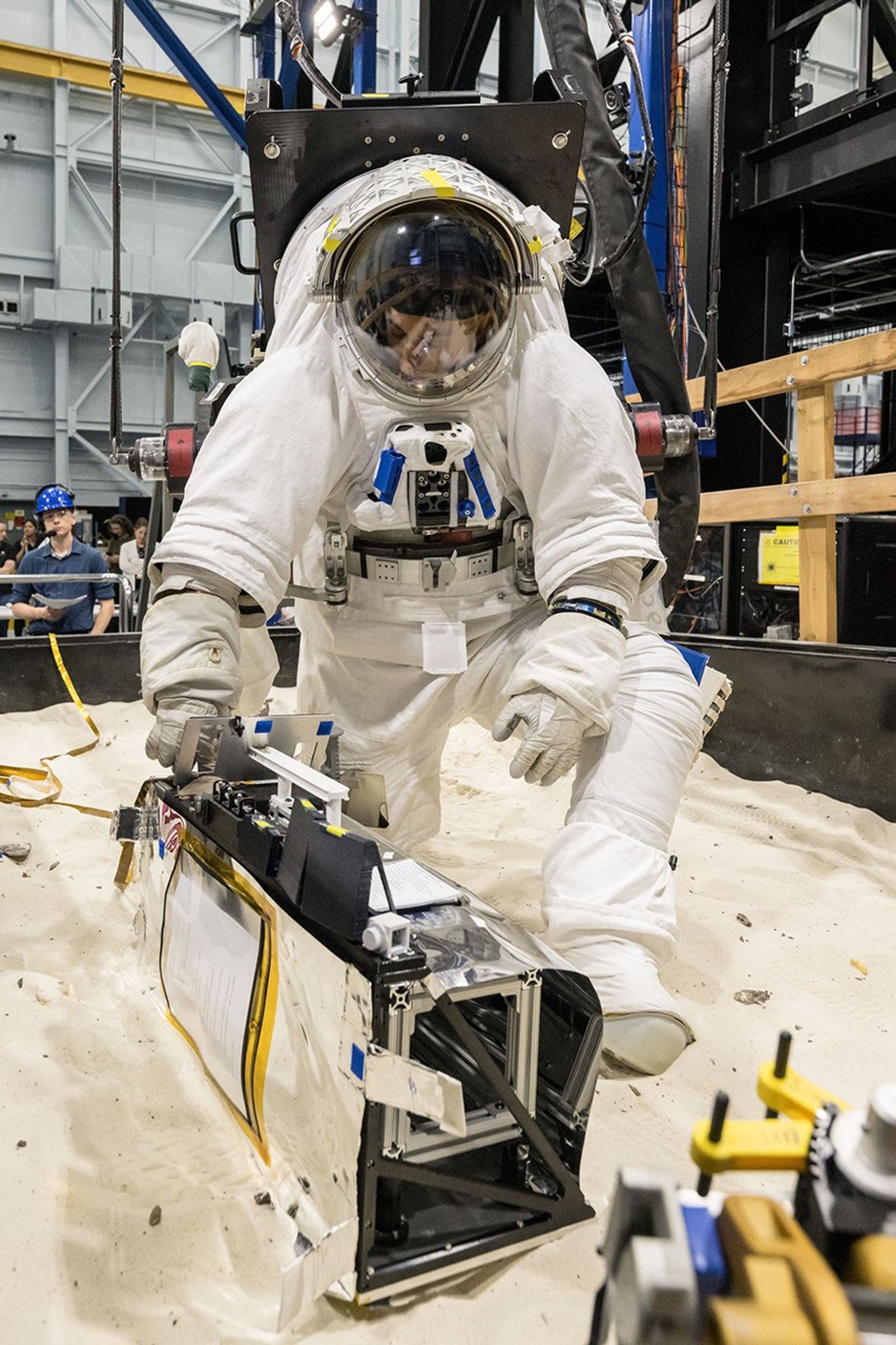 An astronaut in a white spacesuit kneels in simulated lunar soil while working with scientific equipment in a large testing facility. Staff members and support structures are visible in the background.
