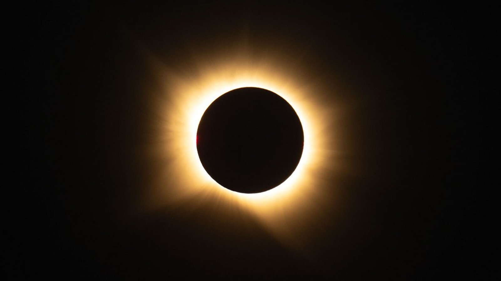 In this photograph of a total solar eclipse, the black disk of the Moon covers the face of the Sun, leaving a glowing, yellow, hazy ring of corona around the edges of the Moon, with large streamers extending toward the upper left, lower left, and lower right.