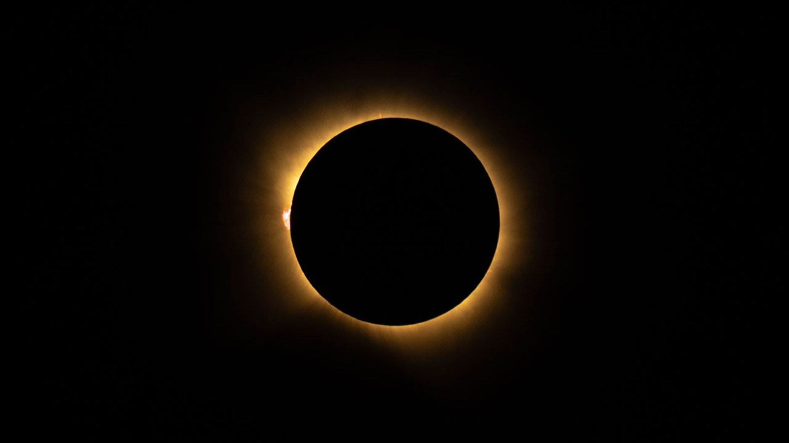 In this photograph of a total solar eclipse, the black disk of the Moon covers the face of the Sun, leaving a glowing, yellow, hazy ring of corona around the edges of the Moon. On the left edge is a pink, mountain-shaped prominence rising off of the edge of the eclipsed Sun.