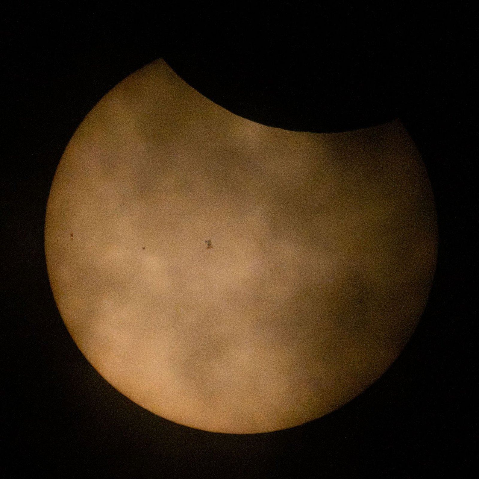 A partially eclipsed Sun appears with the Moon blocking a small portion of the Sun at top. Clouds also appear in front of the Sun. To the left of center is the small silhouette of the International Space Station. A few sunspots appear on the Sun to the left of the space station.