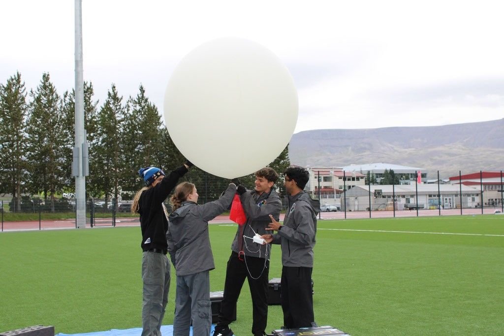 Four students in jackets hold a large white balloon in a grassy field under a gray, cloudy sky.