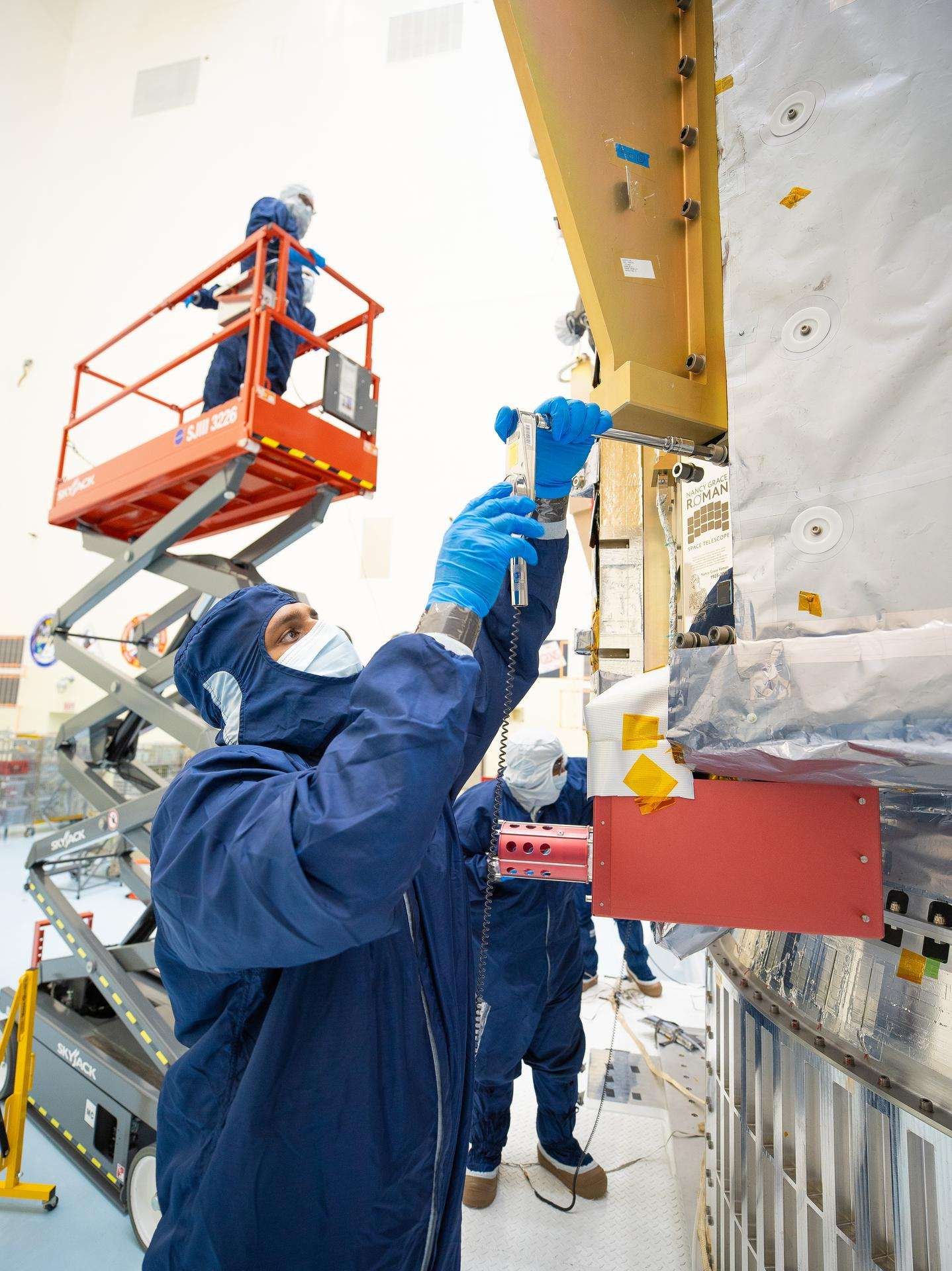 People in blue protective suits and a red cherry picker work in an industrial high bay.