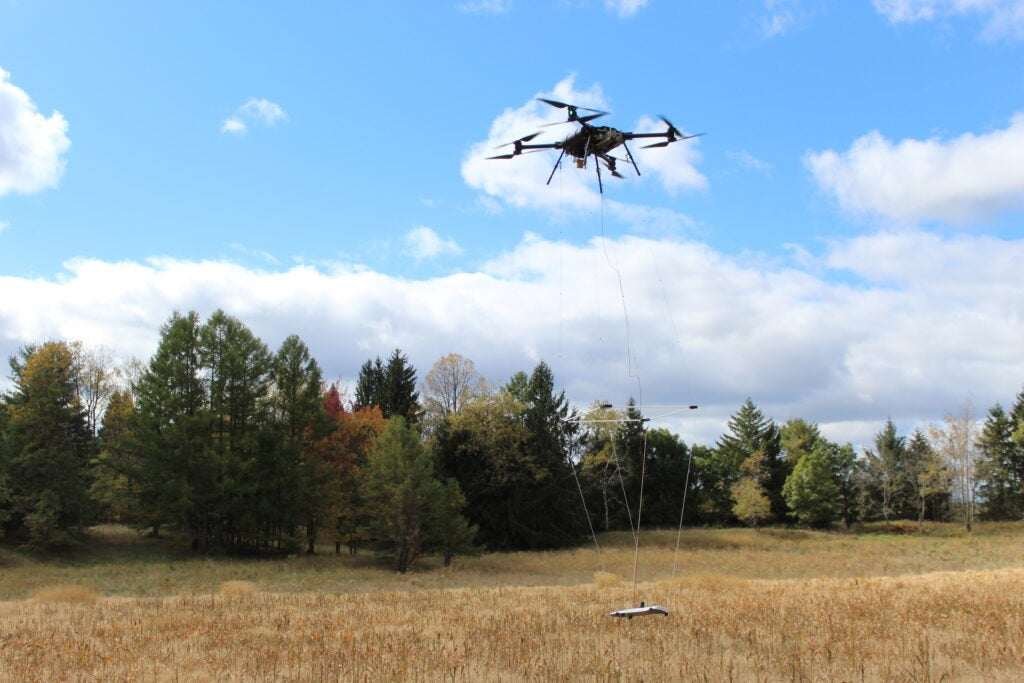 Drone carrying a magnetometer takes flight over a field in Pennsylvania. Autumn treeline is visible in the background against a blue sky and clouds.