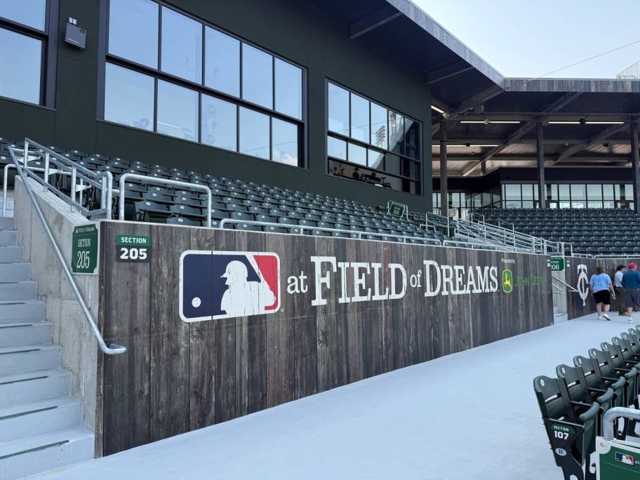 Empty green seats rise behind a weathered wooden wall bearing the MLB at Field of Dreams logo and a John Deere logo, with Section 205 signage. Photo: Dave McEachren