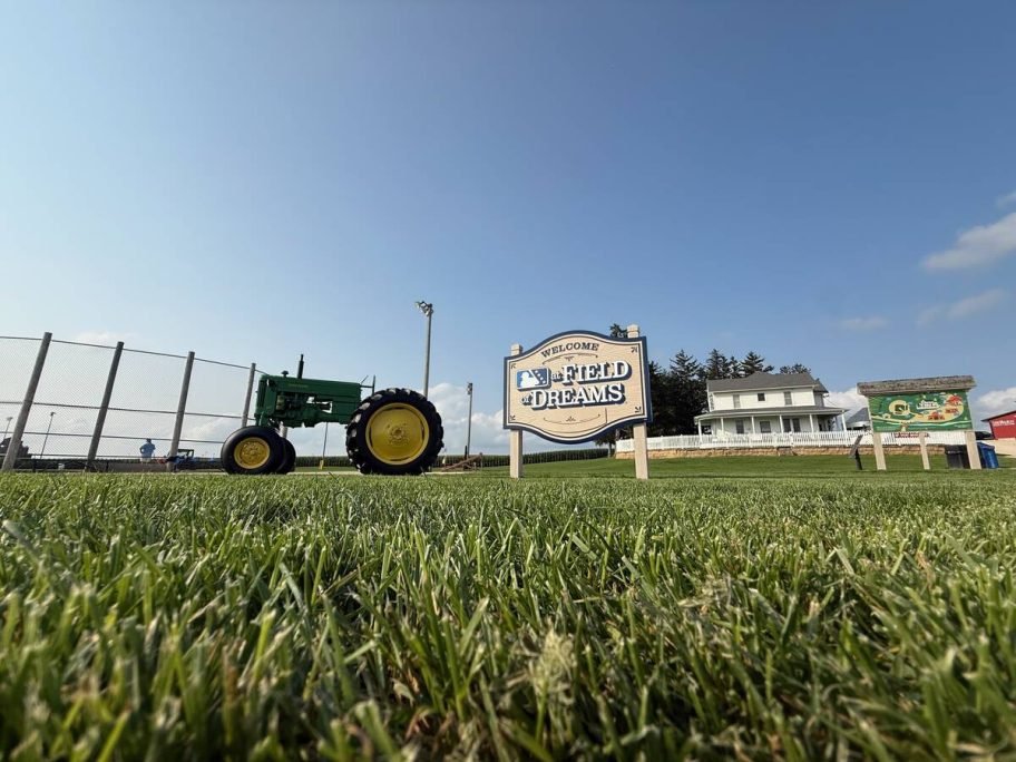 A low-angle view across mown grass showing the John Deere tractor, the Field of Dreams welcome sign and the farmhouse under a clear blue sky. Photo: Dave McEachren