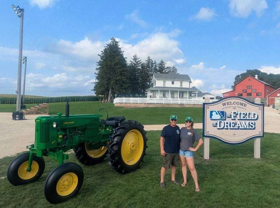 A man and woman in matching ball caps stand together between the green John Deere tractor and the Field of Dreams welcome sign. Photo: Dave McEachren