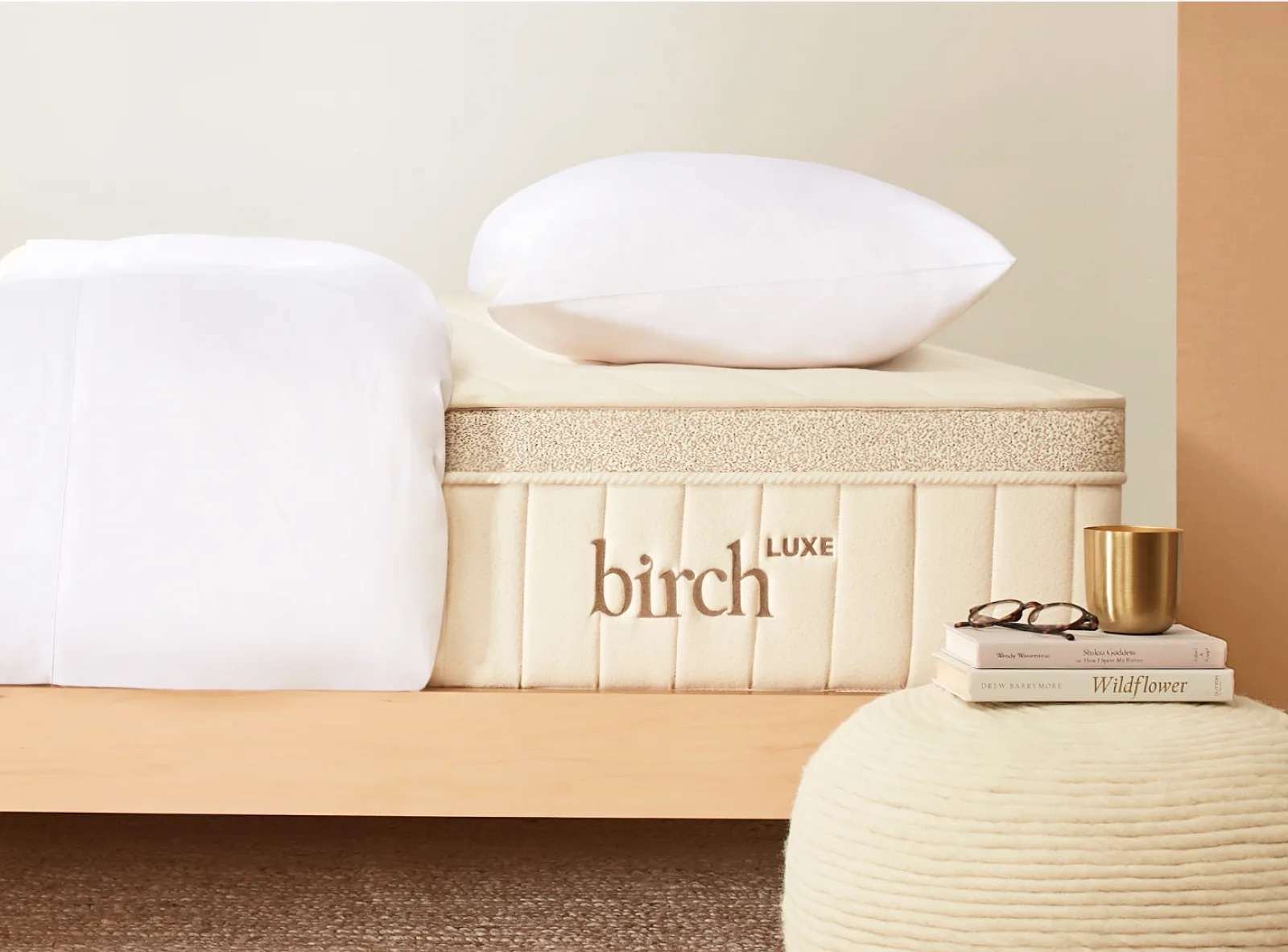Mattress with "Birch Luxe" embroidered, topped with white bedding and a pillow. Nearby are a white pouf, a stack of books, and a gold cup.