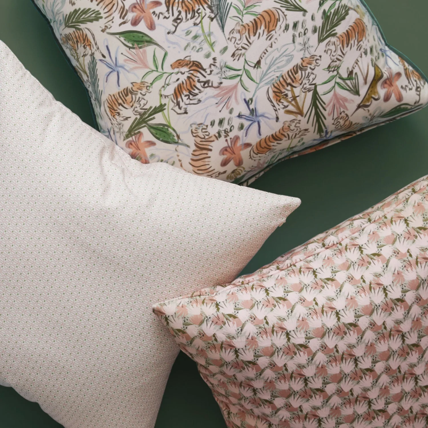 Three decorative pillows on a green surface, one with a tiger and floral print, one with a small dotted pattern, and one with a pink and green leafy design.