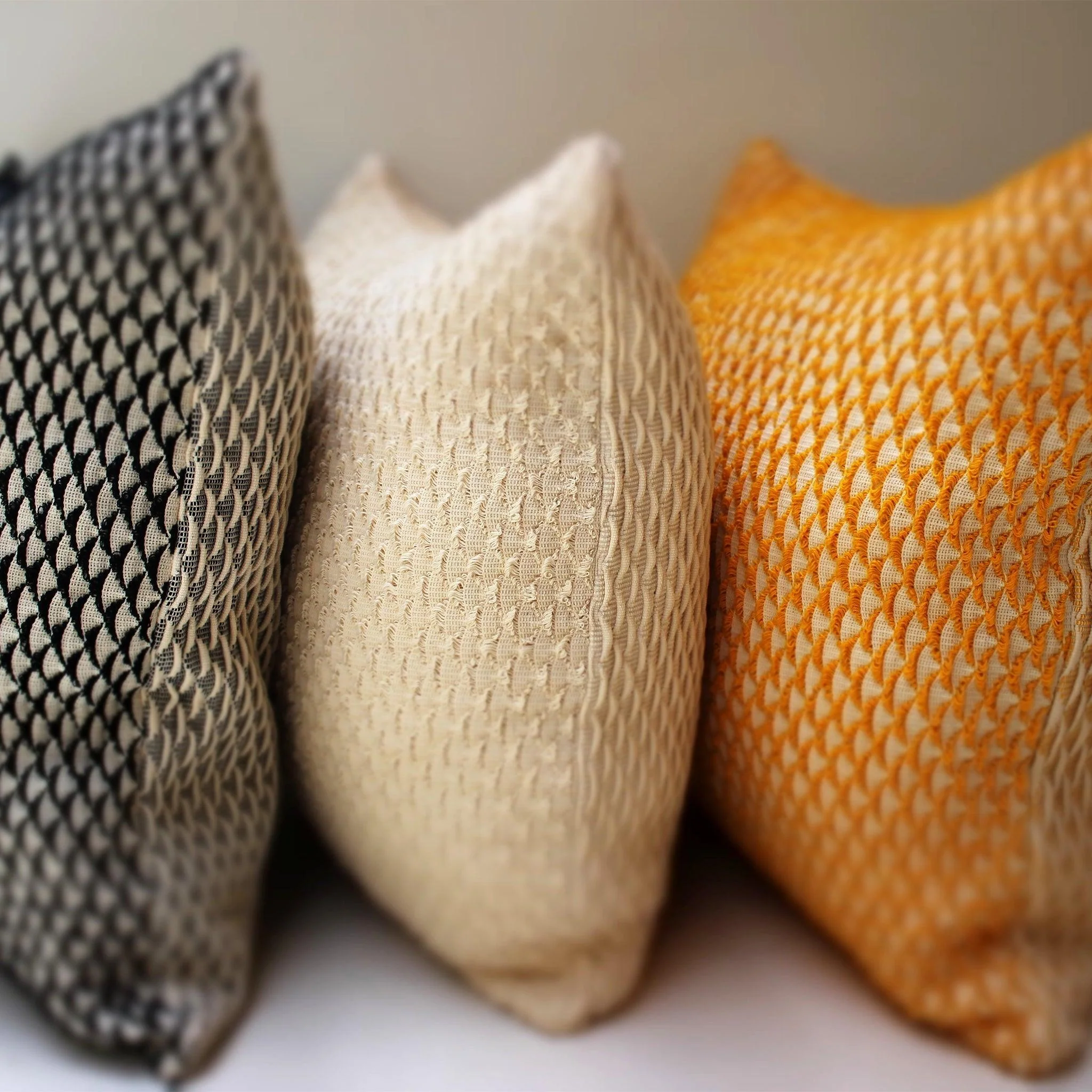 Three textured decorative pillows in gray, off-white, and yellow are arranged side by side against a neutral background.
