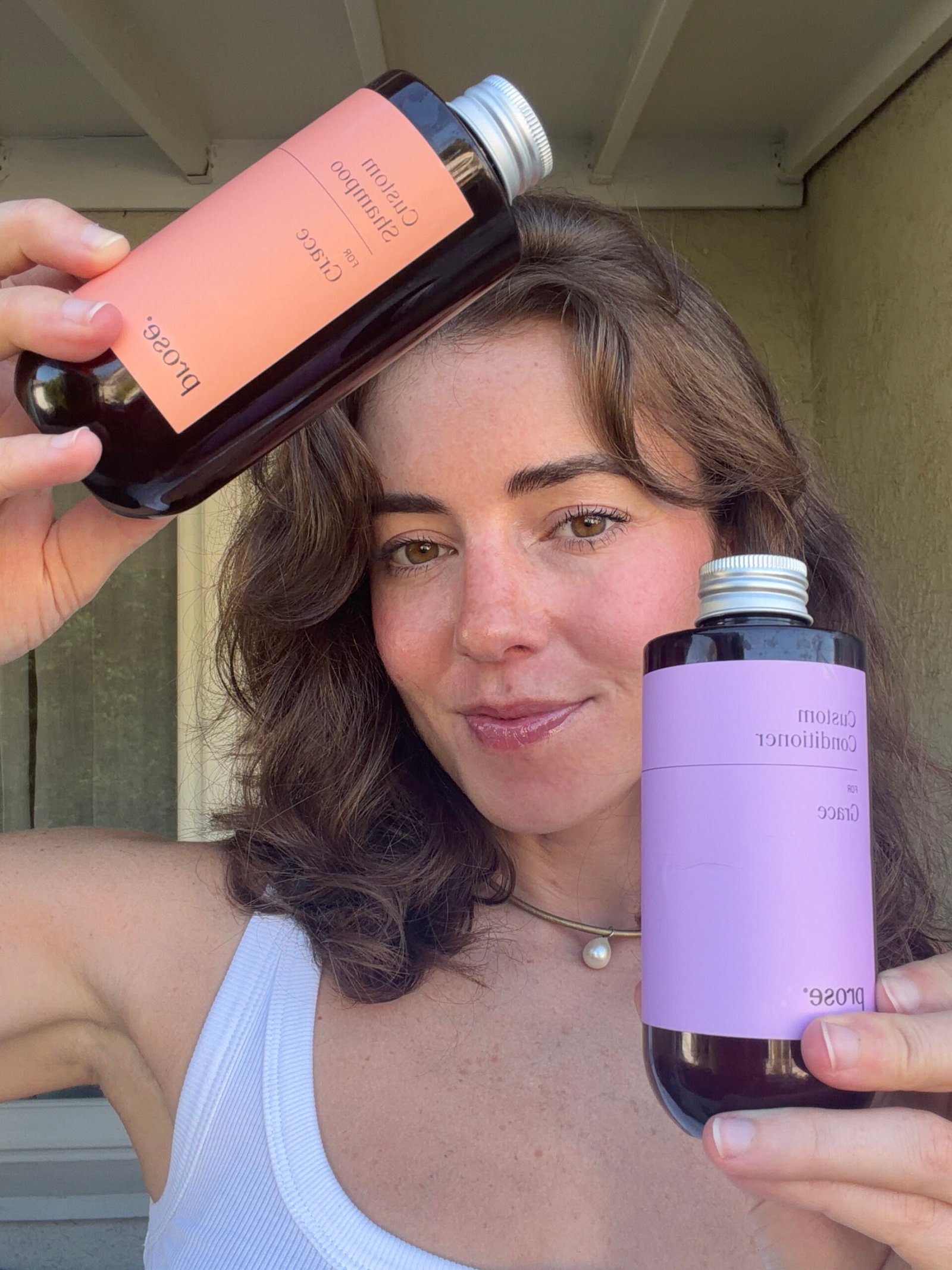 Woman in a white tank top holds two bottles labeled “Custom Shampoo” and “Custom Conditioner” by Prose while standing outside.