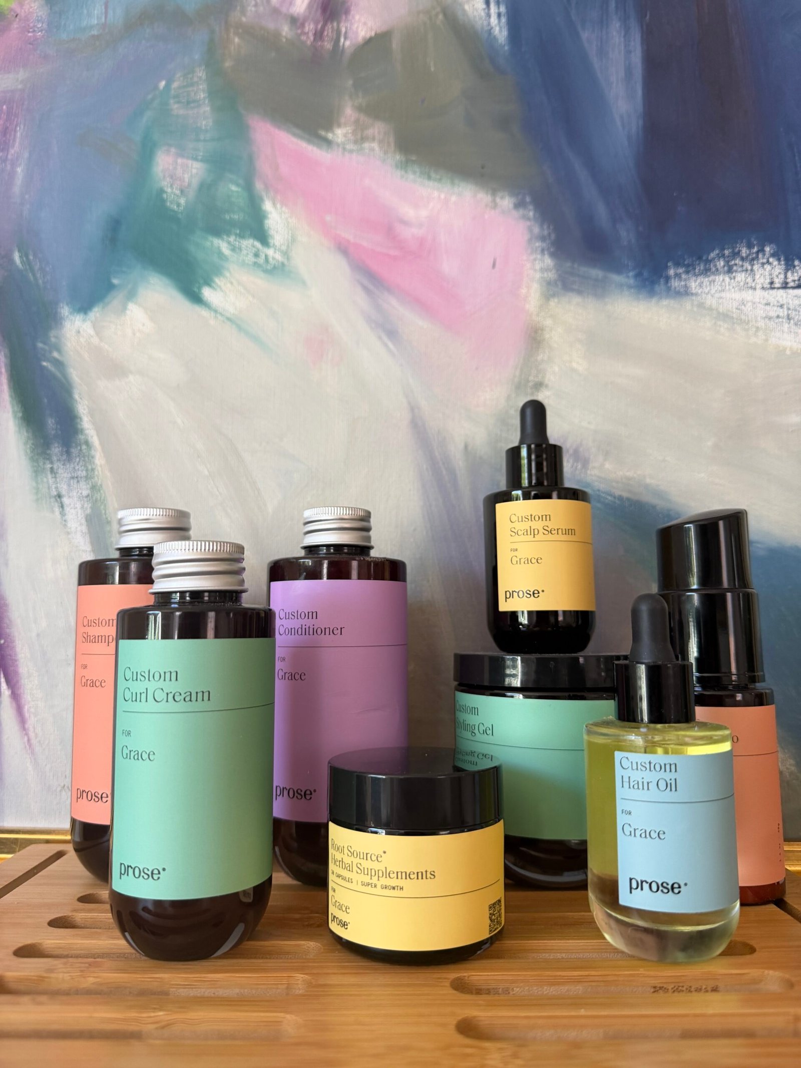 Various custom-labeled Prose hair and skin products, including bottles, jars, and dropper vials, are arranged on a wooden tray against an abstract colorful background.