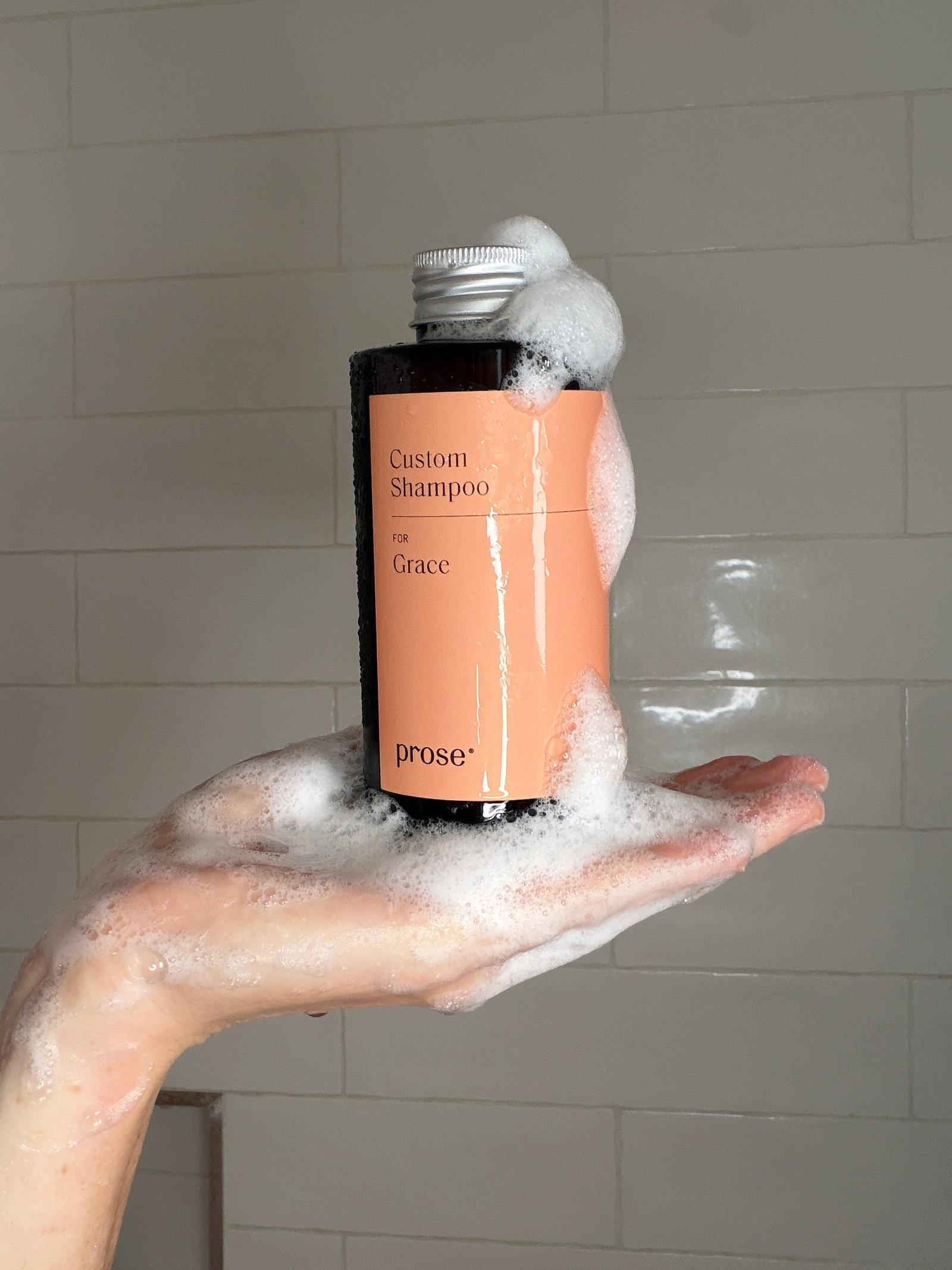 A hand covered in soap suds holds a bottle labeled “Custom Shampoo for Grace” by Prose against a tiled shower wall.